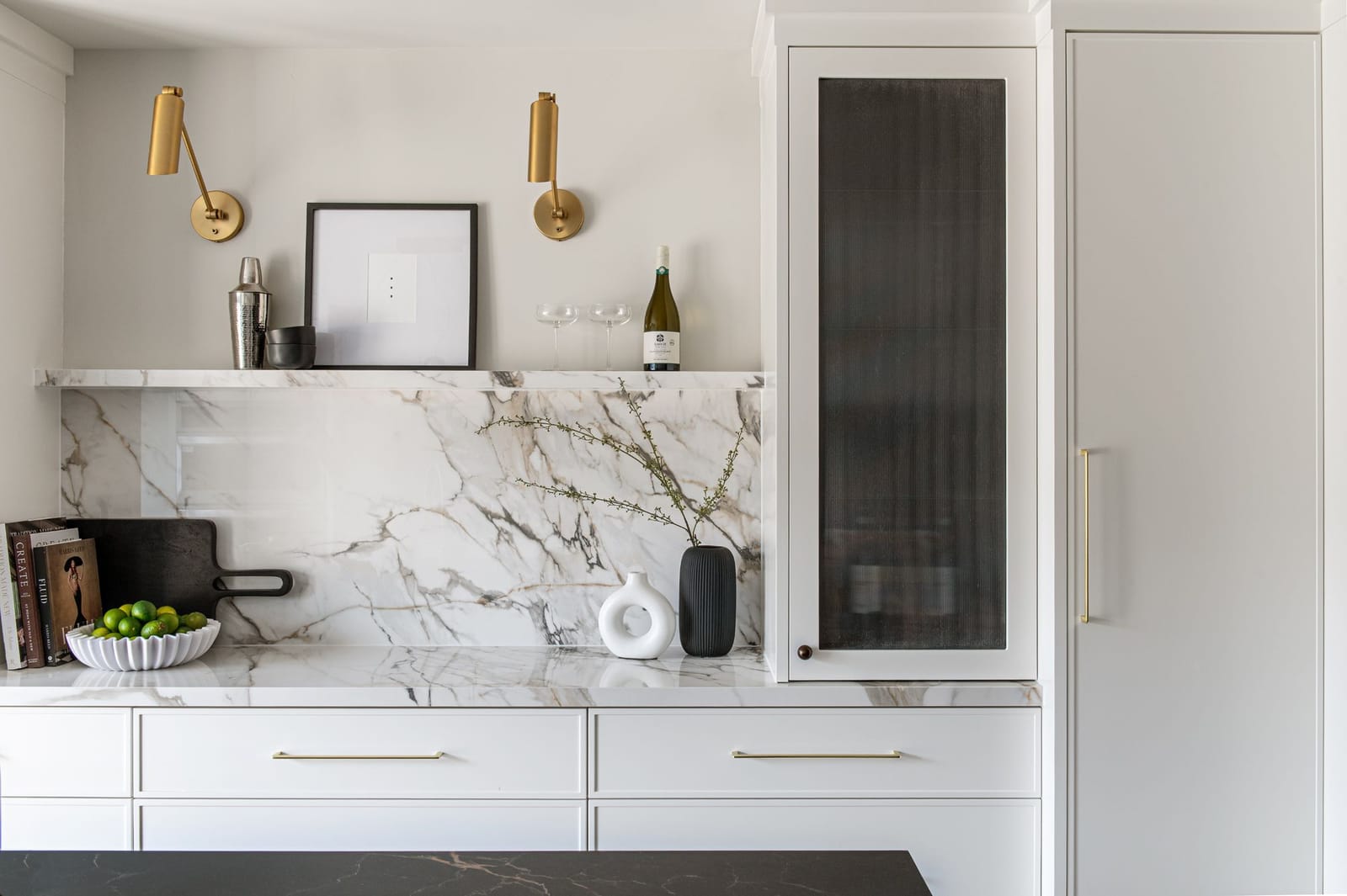 A bar wall with a white and grey-veined marble backsplash and shelf, two brass swing-arm lights, and tall cabinets with reeded black glass