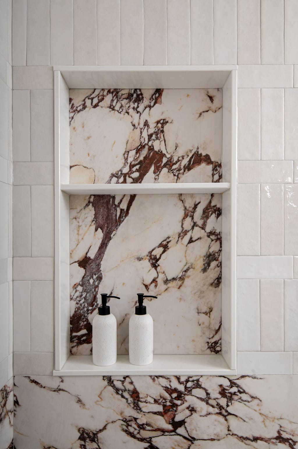 A shower niche lined in white marble with dark red veining, framed by glossy white vertical tile
