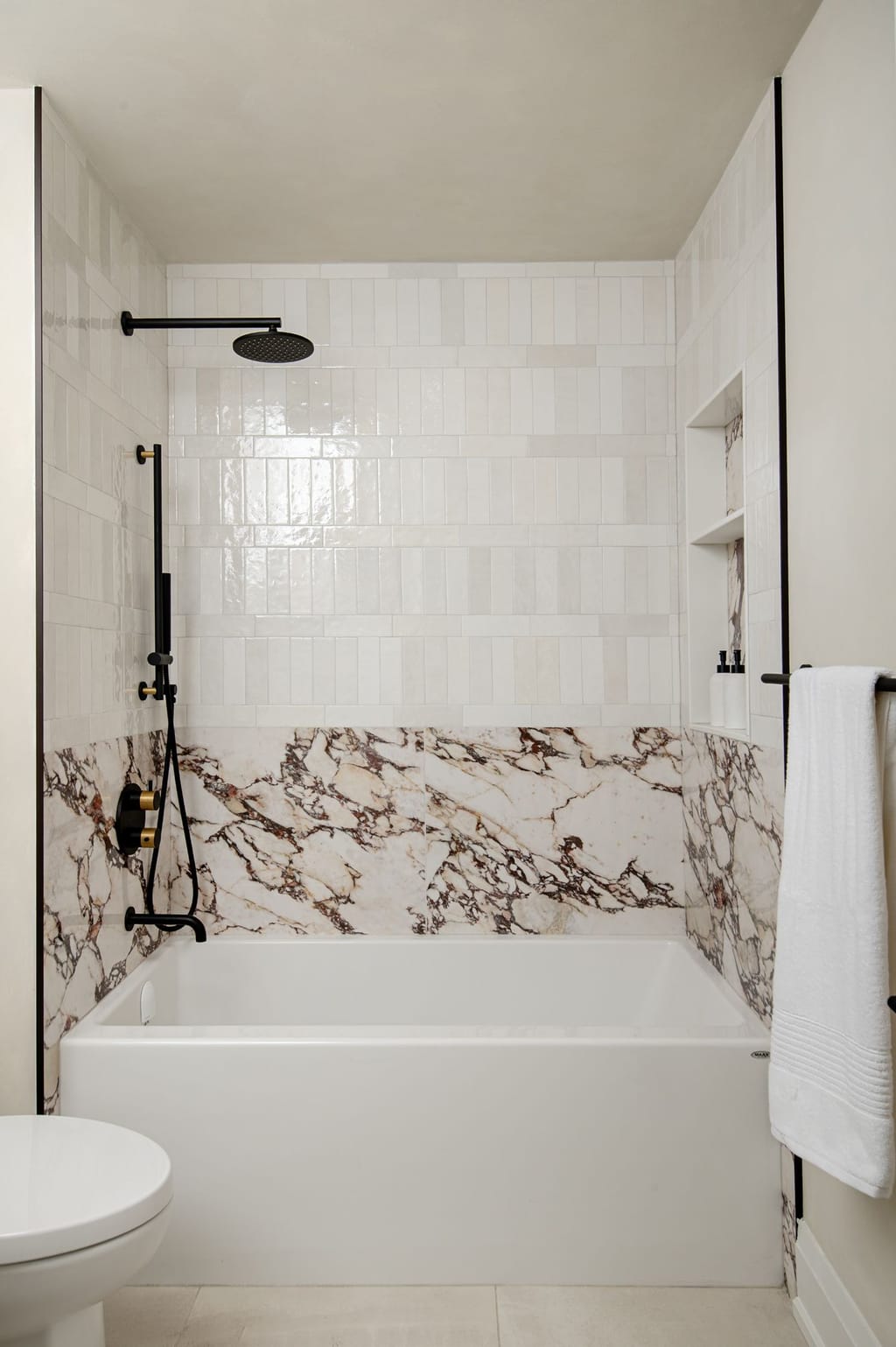 A tub alcove tiled in glossy white vertical tile above a band of white marble with dark red veining, with black shower fittings