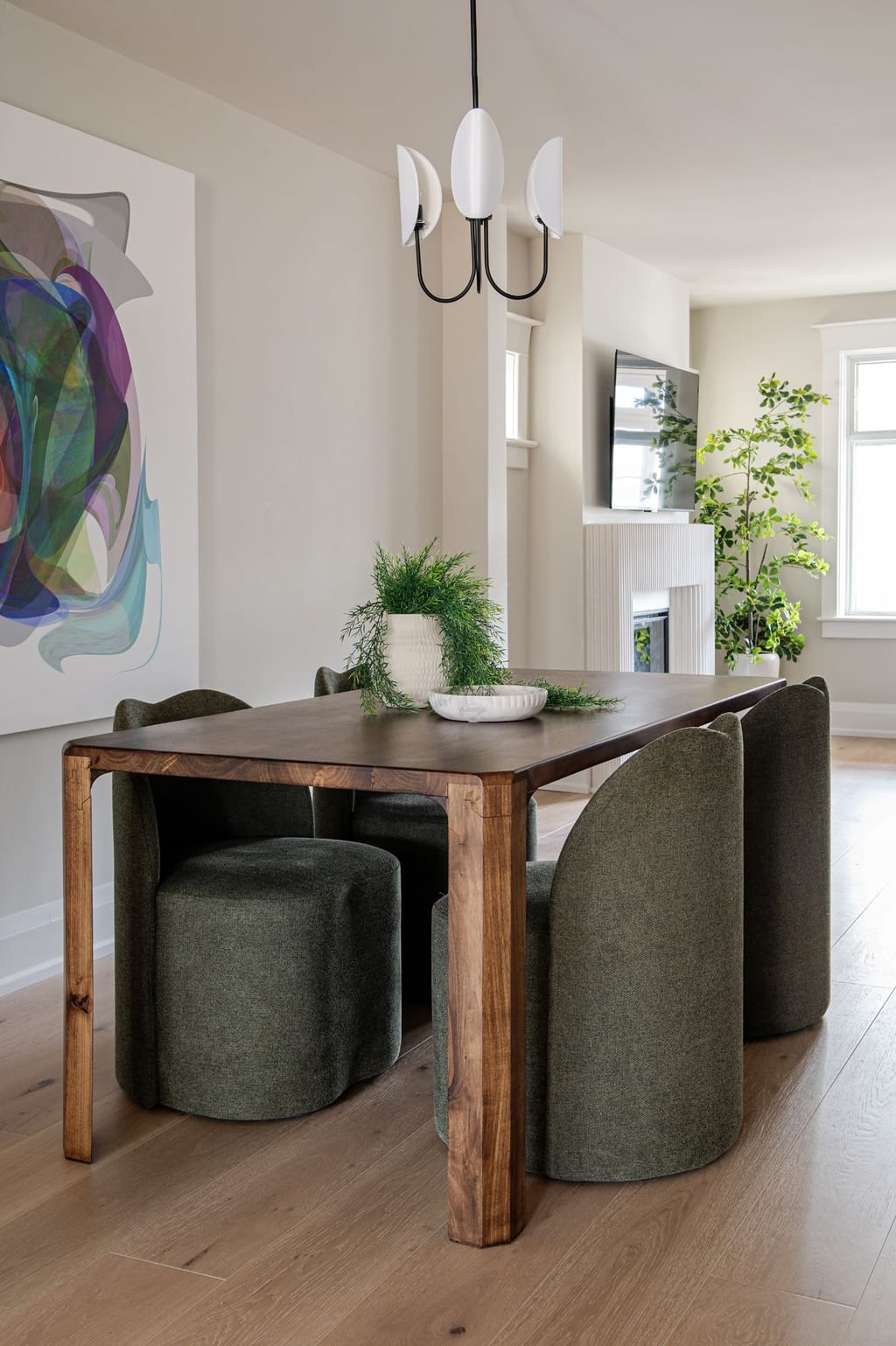 A dining table in walnut with green upholstered chairs, an abstract painting and a three-shade pendant, looking through to the living room