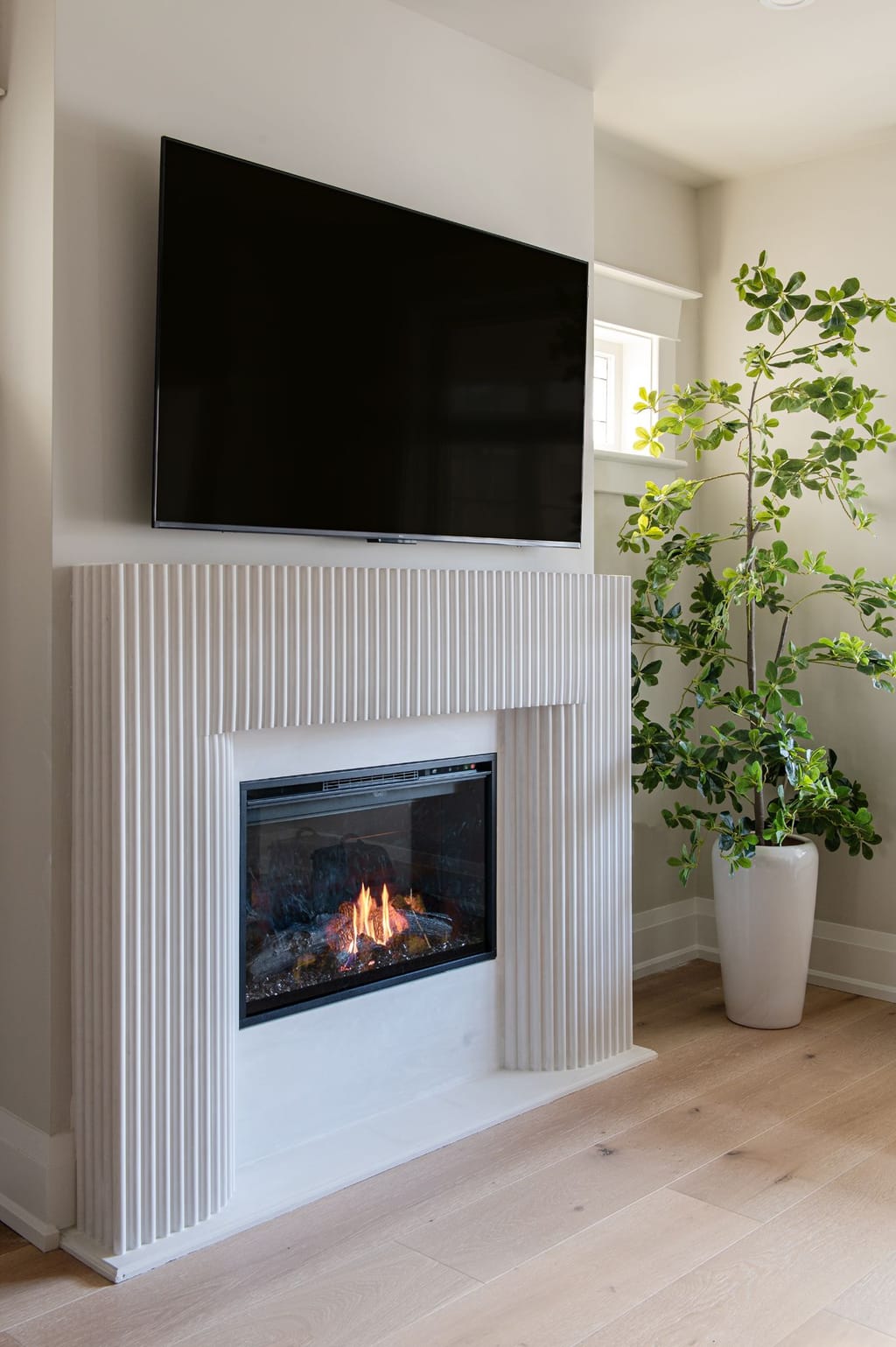 A white fluted fireplace surround around a gas insert, with a television above and a small leaded window to the side