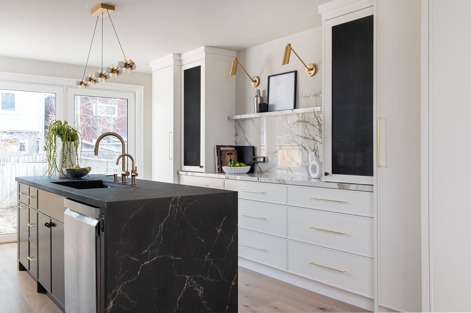 A long kitchen with a black porcelain waterfall island veined in gold, white flat-panel cabinets, reeded glass uppers and doors to the garden