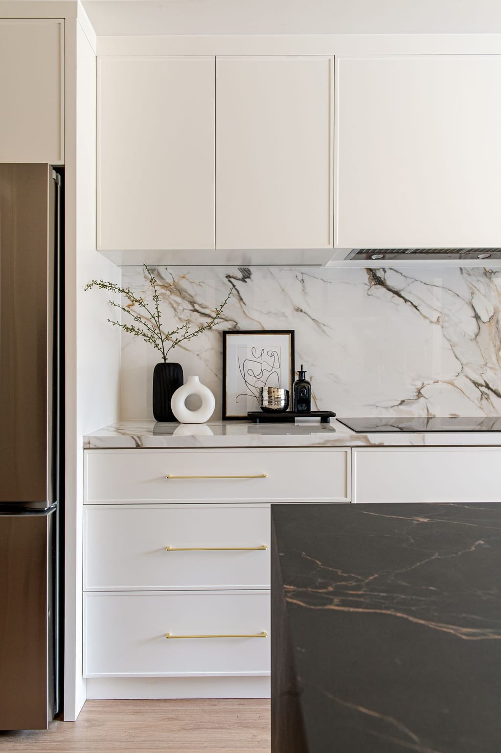 White slab cabinets with brass pulls and a marble backsplash behind a cooktop, with the corner of the black waterfall island in front