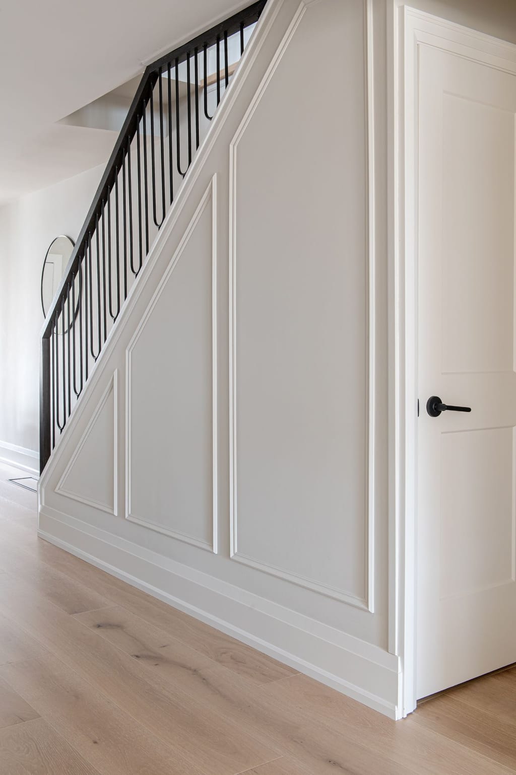 The underside of the stair clad in white picture-frame panelling beside a white door with a black handle