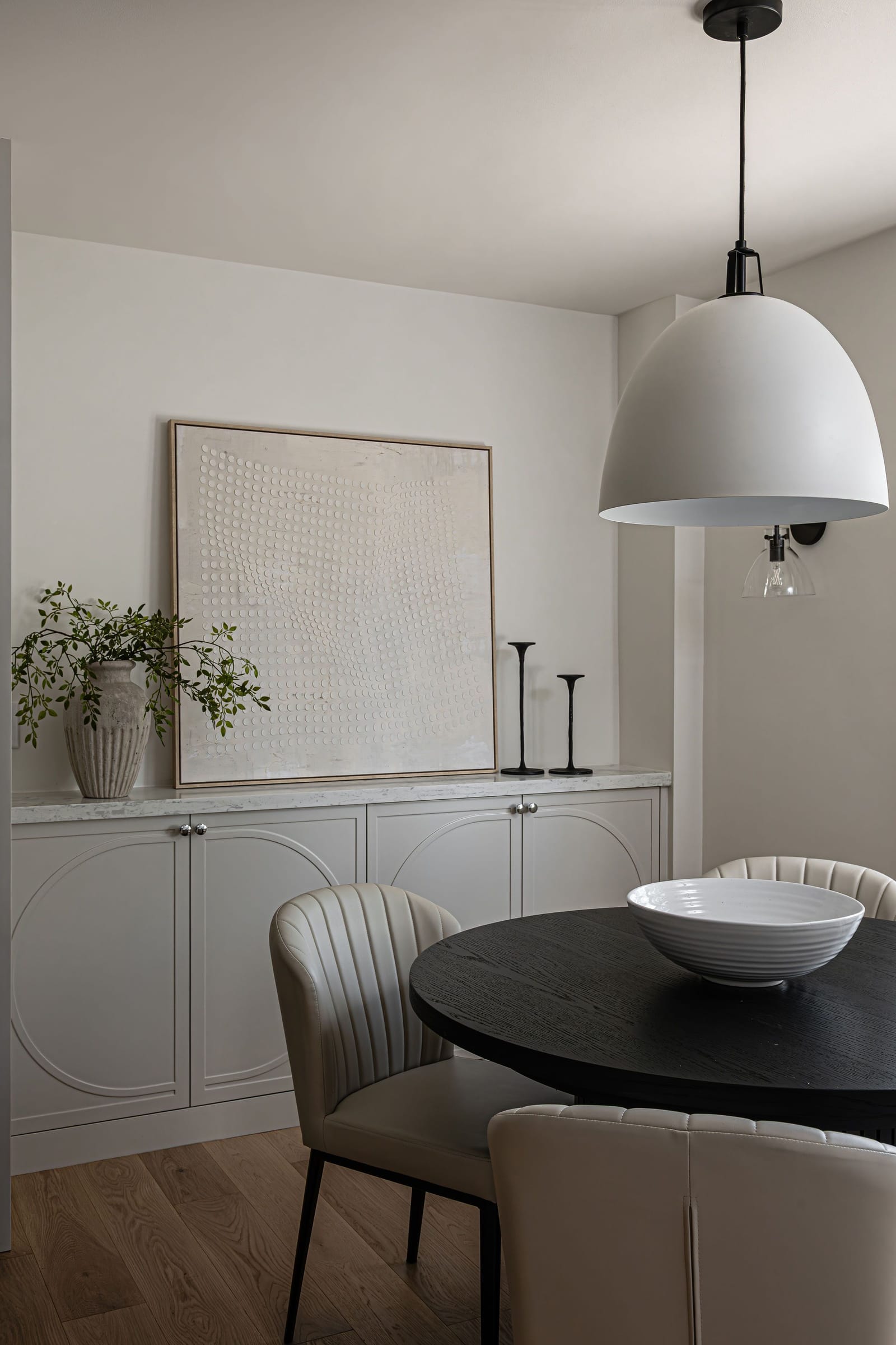 Cream sideboard cabinets with arched panel doors under a textured artwork, beside the round black dining table and a white dome pendant