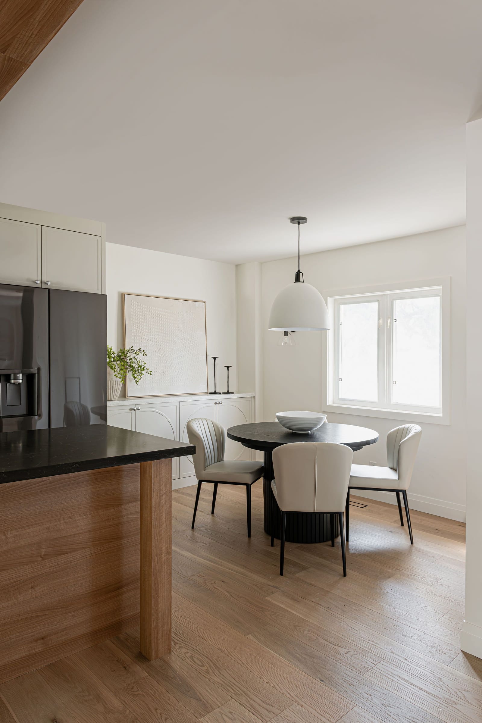 View past the end of the island to a dining nook with a round black table, cream curved chairs and a white dome pendant by the window
