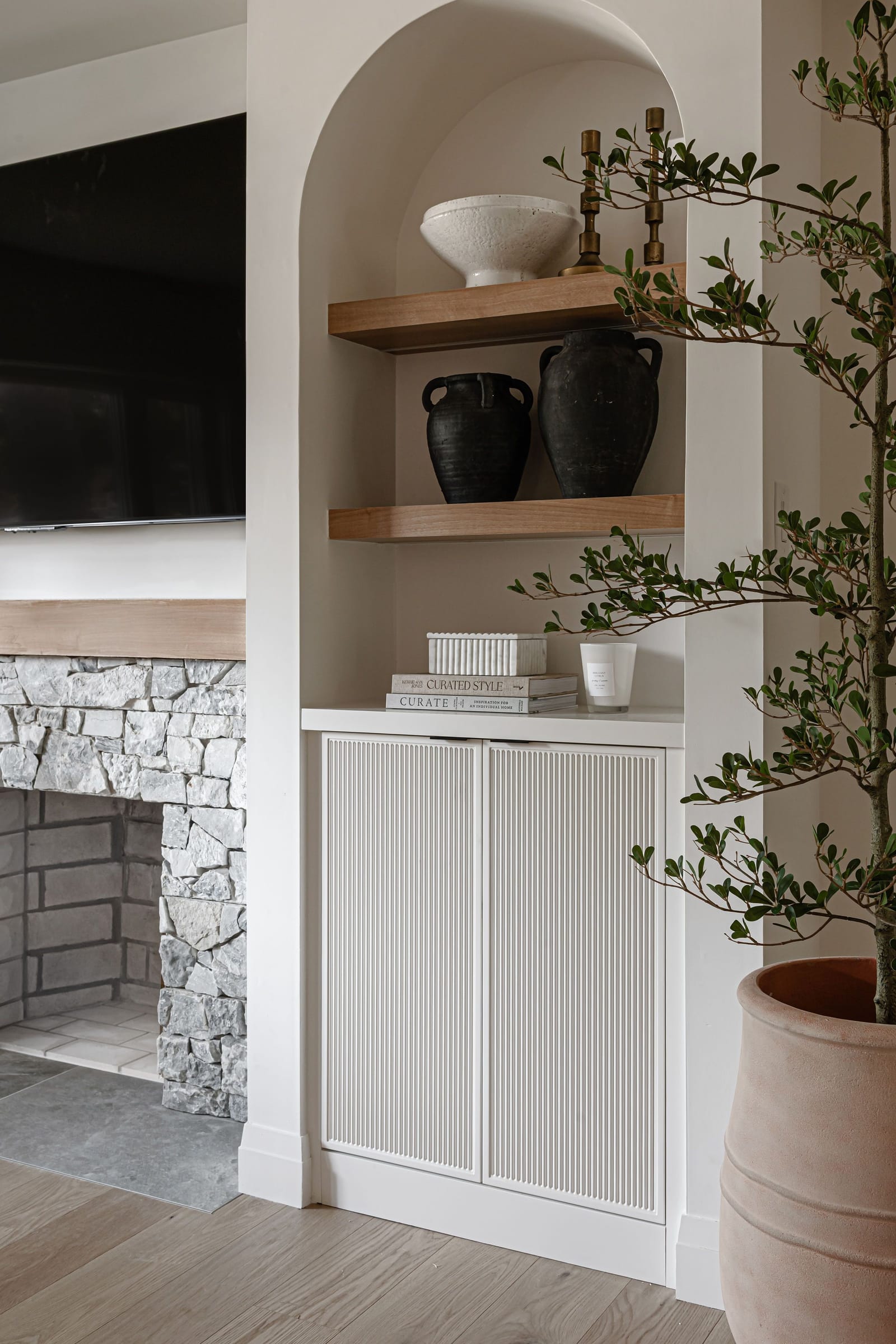 An arched built-in with oak shelves and fluted white doors beside a stacked natural stone fireplace with an oak mantel