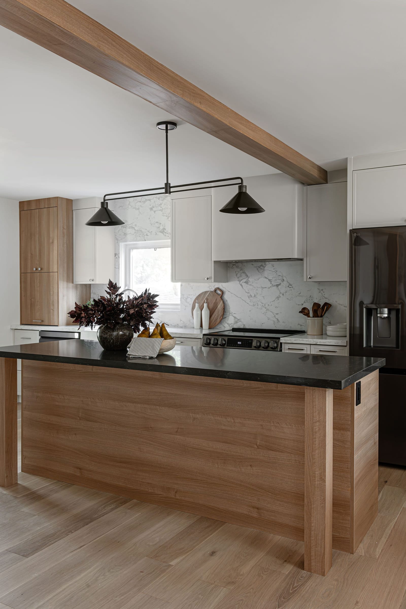 A long island clad in pale oak with a dark stone top and squared legs, under a black two-shade pendant and an oak-wrapped beam