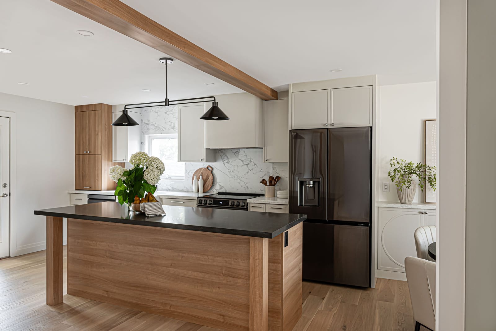 An open kitchen in a brick bungalow with an oak-wrapped beam across the ceiling, a wood-sided island with a black top and a dark French-door fridge