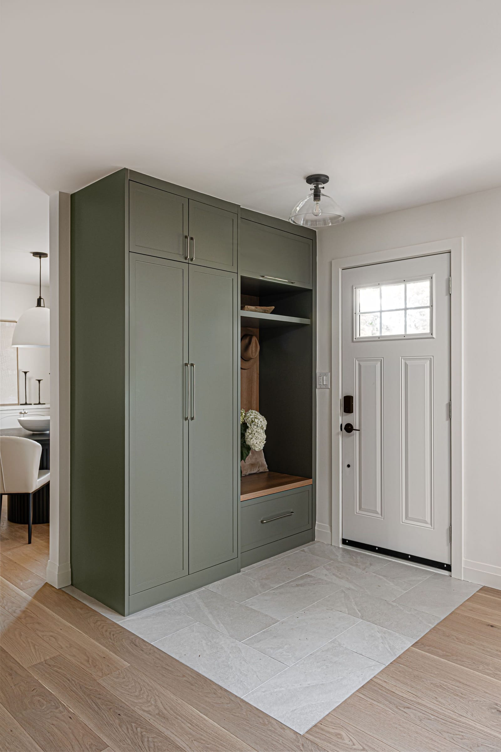 A sage-green mudroom wall unit with tall cabinets, coat hooks and a bench, on a grey tile floor beside the front door
