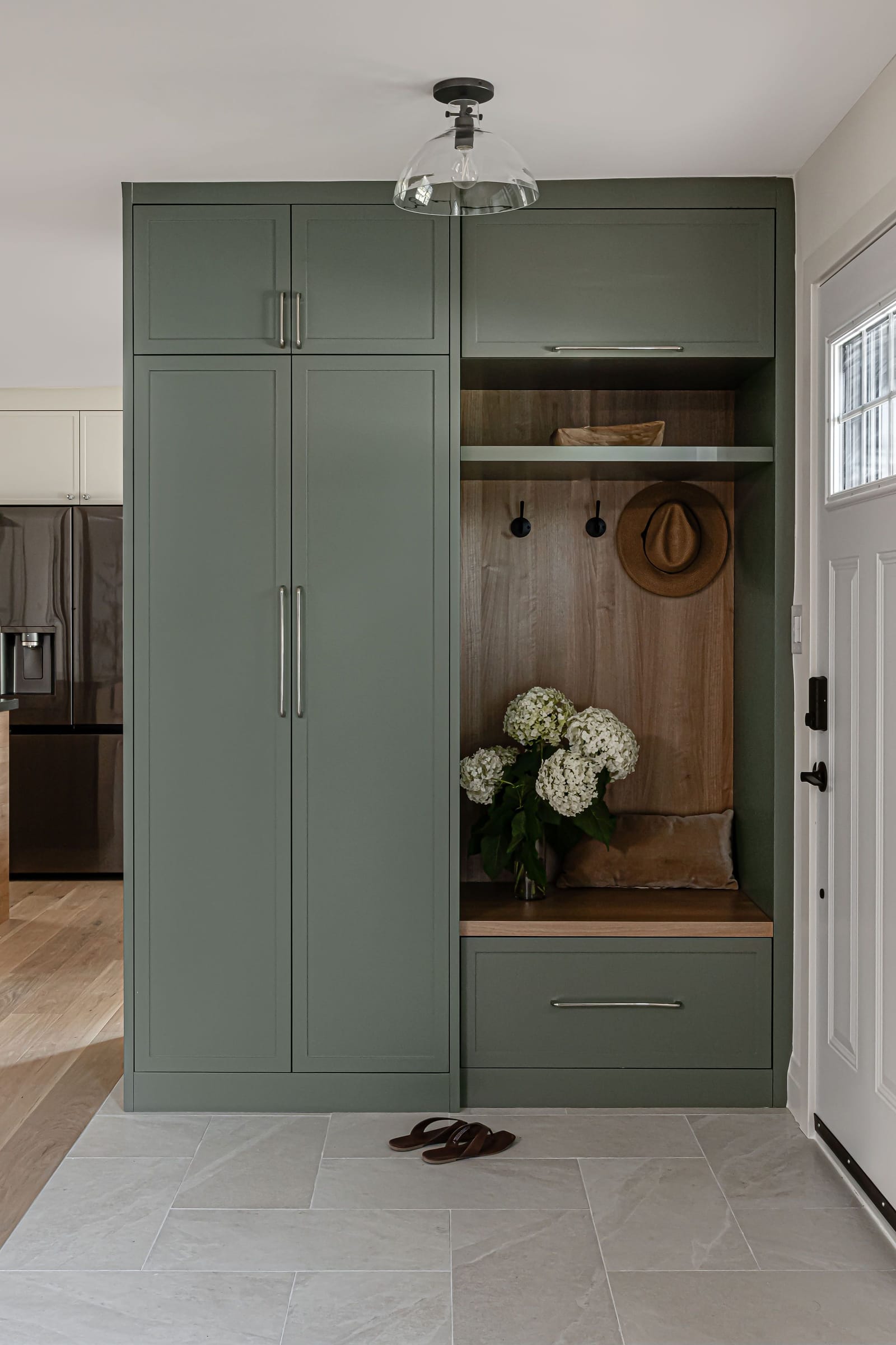 Straight-on view of the sage-green mudroom unit with an oak-lined bench, hooks, a hat and sandals on the tile