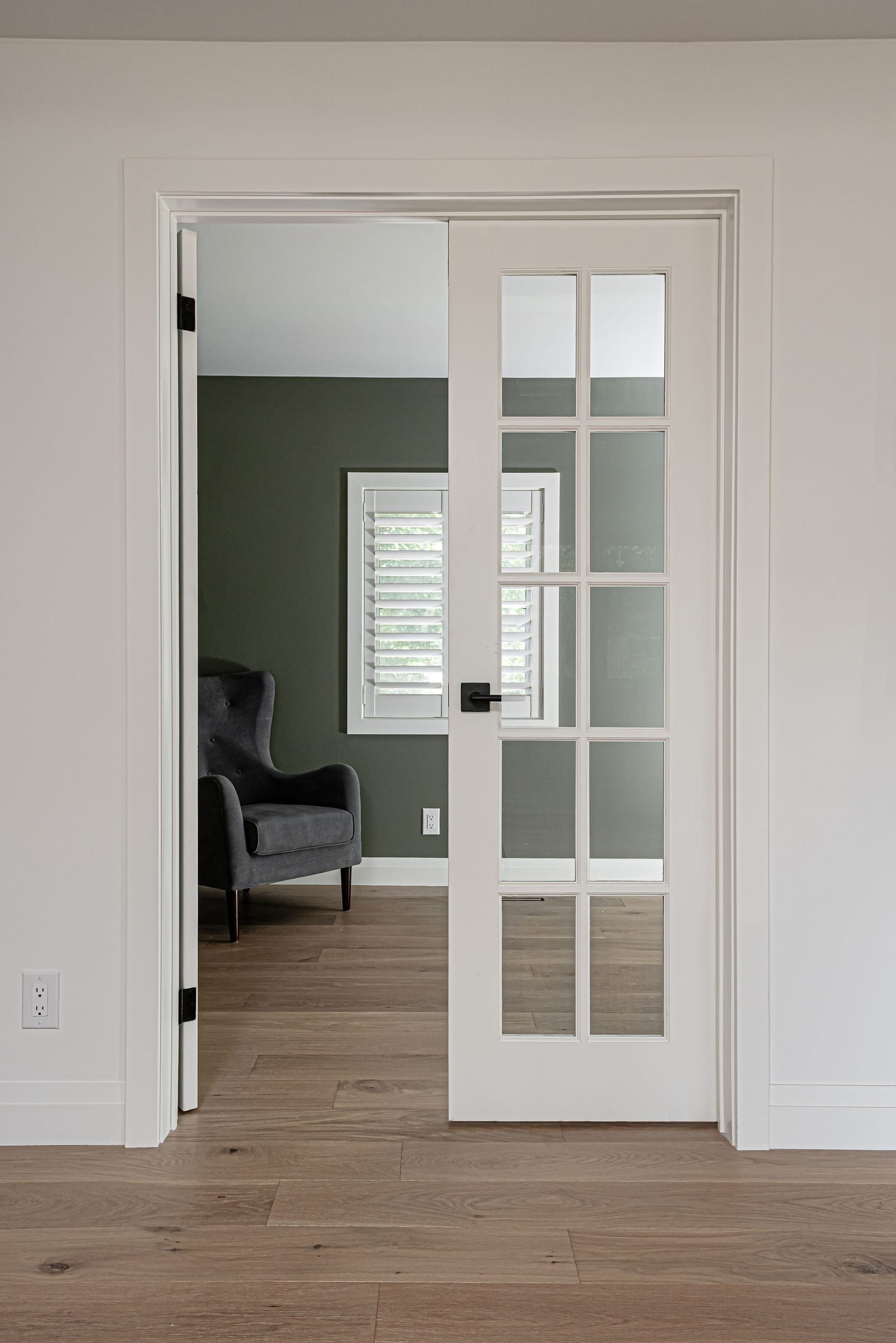 White French doors with glass panes opening onto an office painted dark green, with a grey armchair by a shuttered window