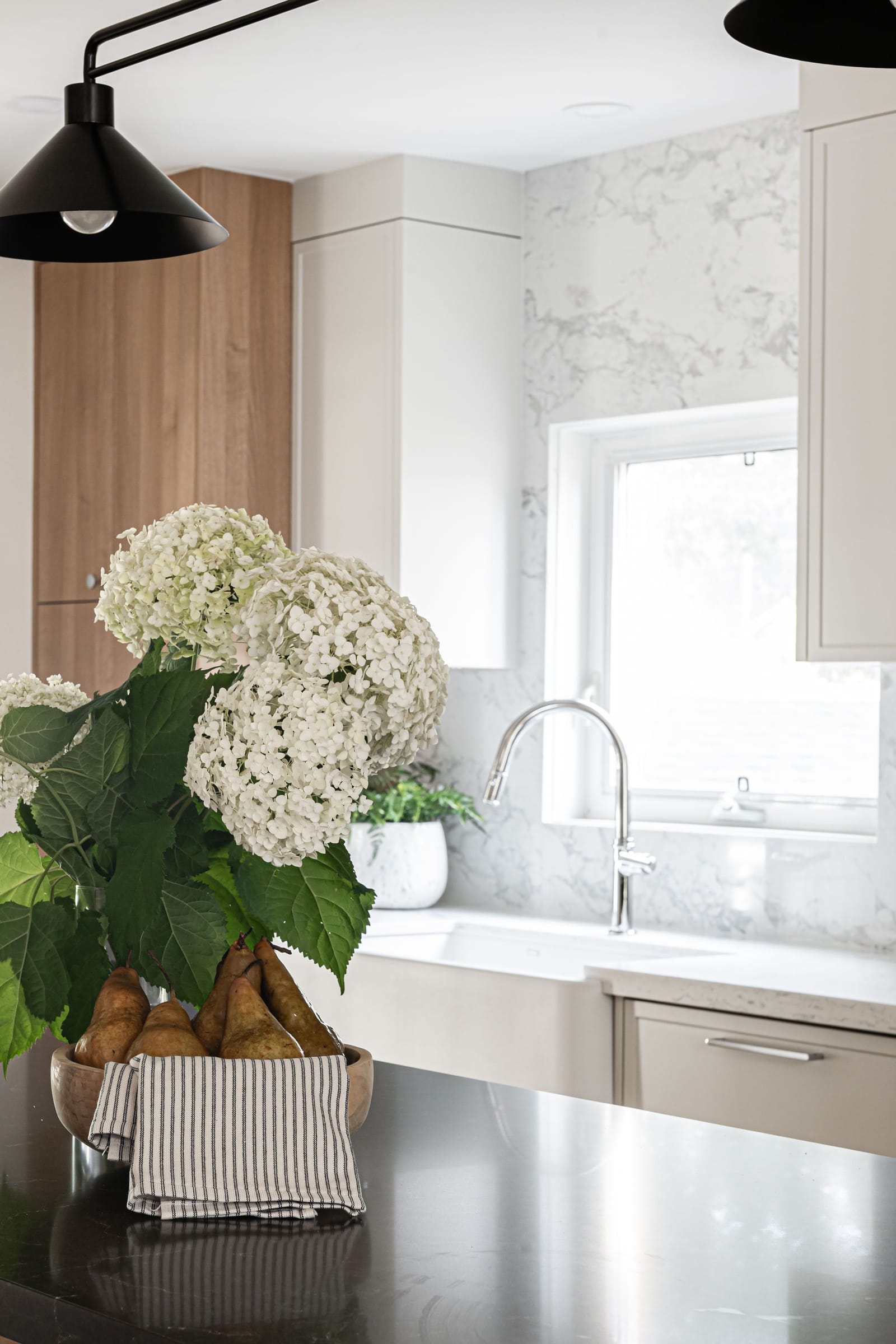A white farmhouse sink with a chrome faucet under a window, behind the black island top and a vase of white hydrangeas