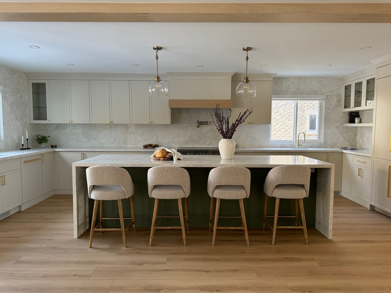 A kitchen with a waterfall island in cream veined stone over sage-green cabinets, four stools and cream cabinets