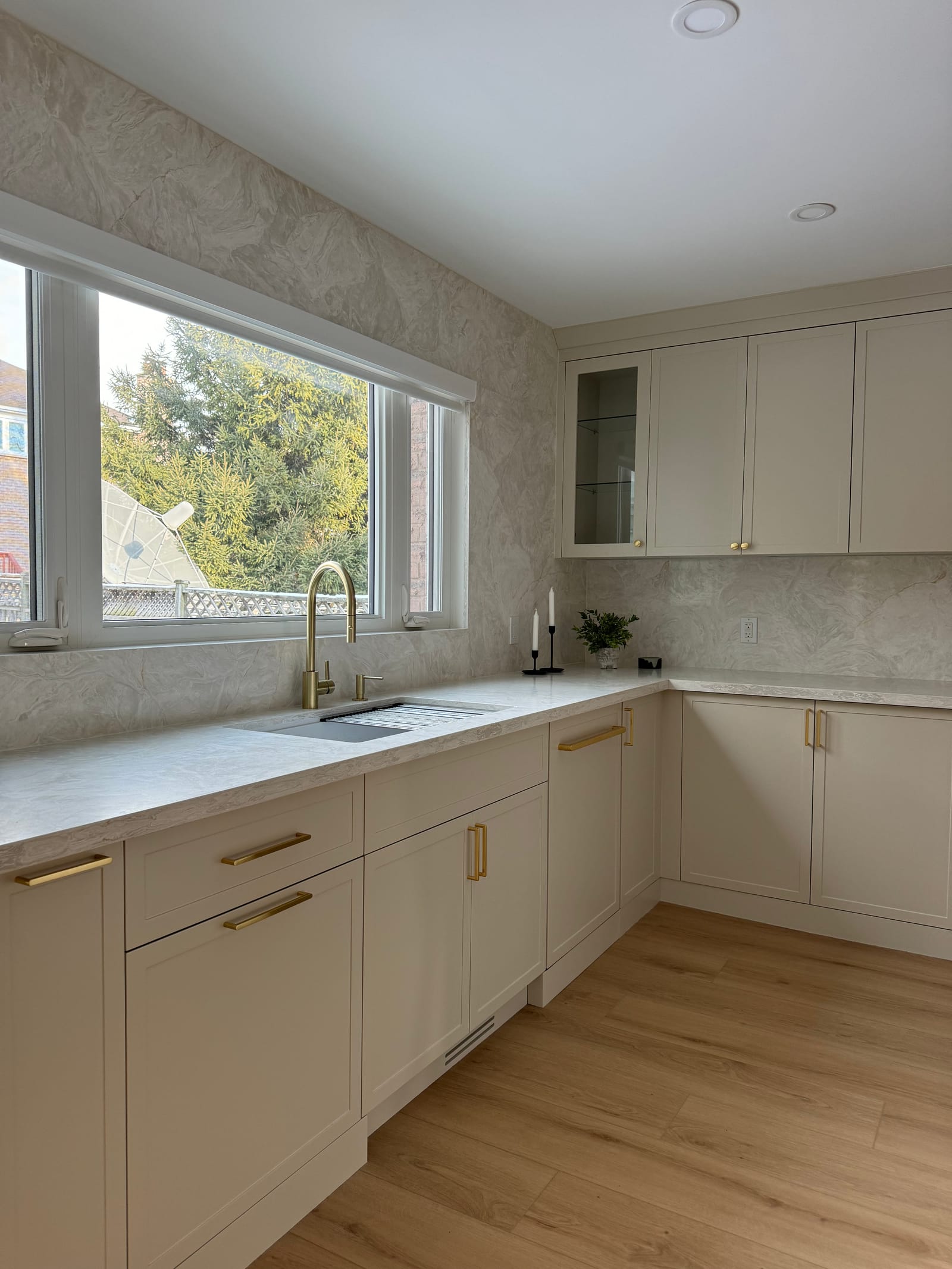A cream sink run under a wide window, with a stone backsplash and brass hardware