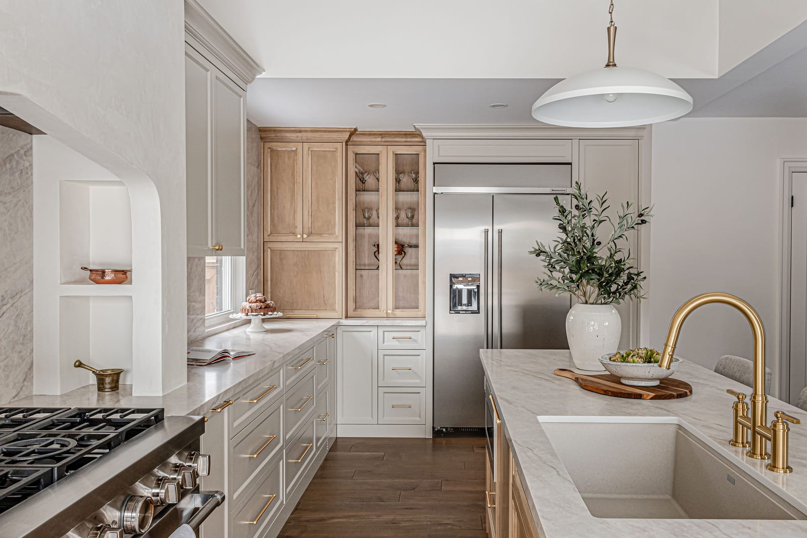 A brass faucet on a stone-topped island, with the range, cream cabinets and maple glass-front cabinets beyond