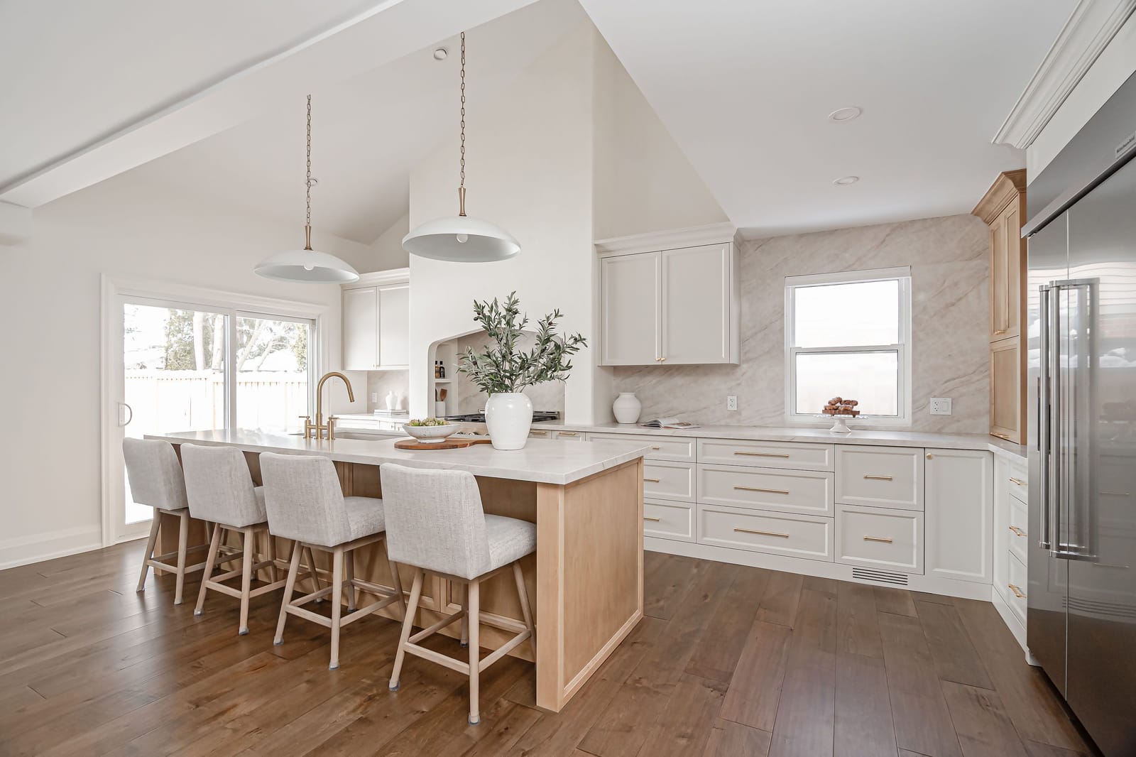 A kitchen with a vaulted ceiling, a long white oak island with four stools, cream cabinets and a plaster range alcove