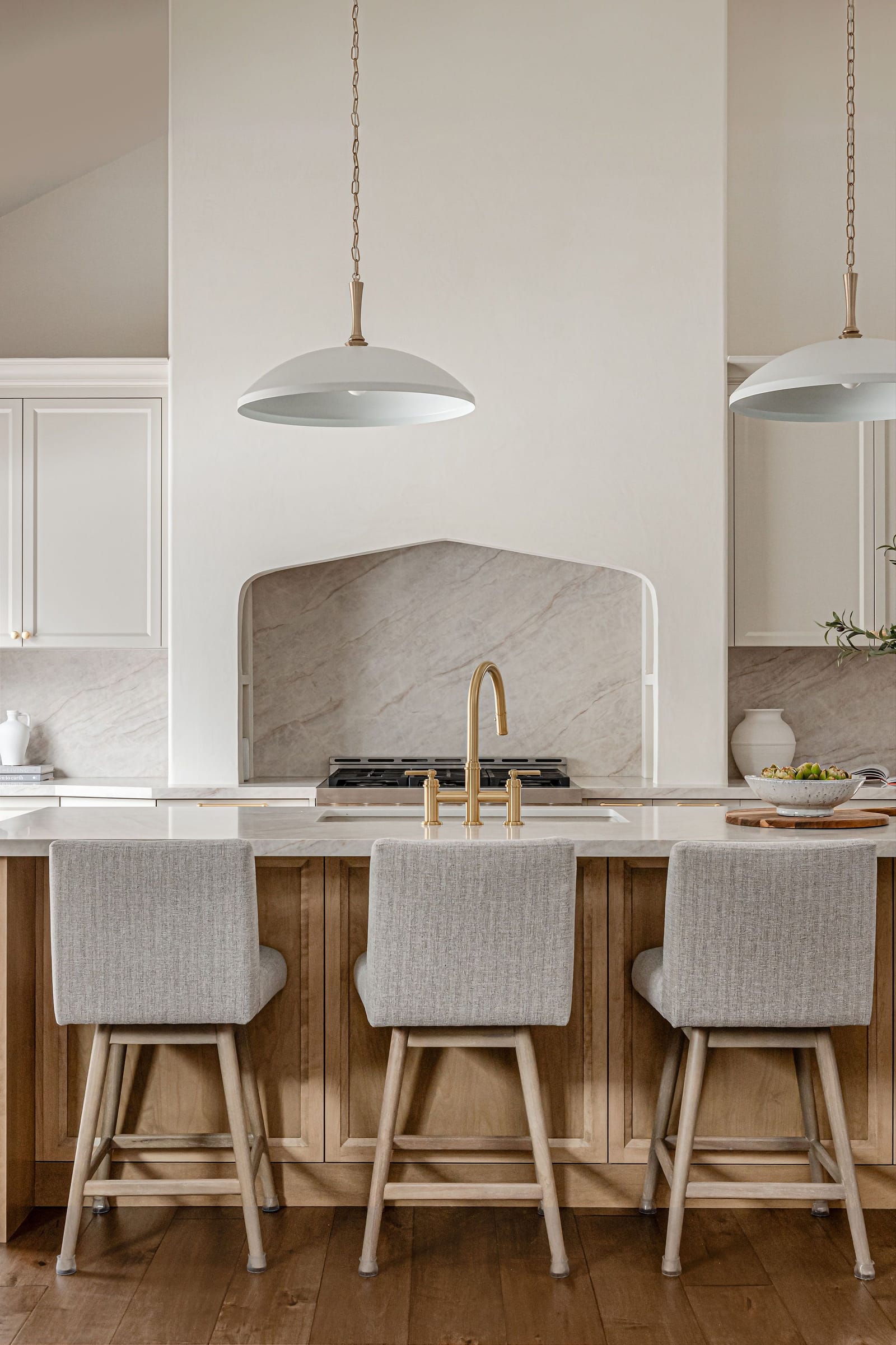 A plaster range alcove with a stone slab backsplash behind a gas range, seen across a white oak island