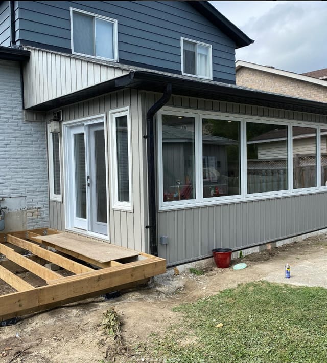 A single-storey rear addition in grey vertical siding with wide windows and patio doors, with deck framing in front