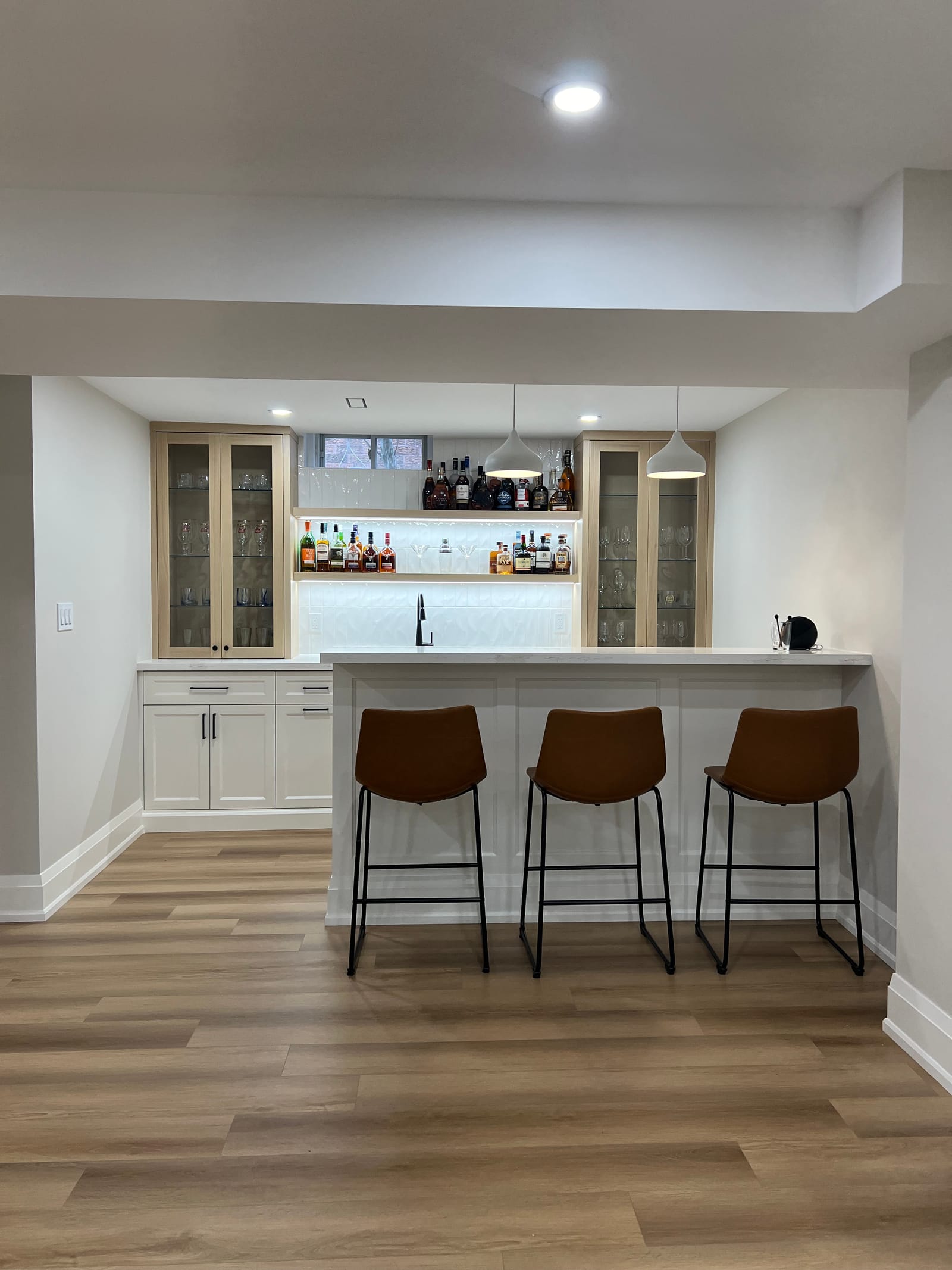A basement bar with a white peninsula, three leather stools, glass-front cabinets and lit bottle shelves
