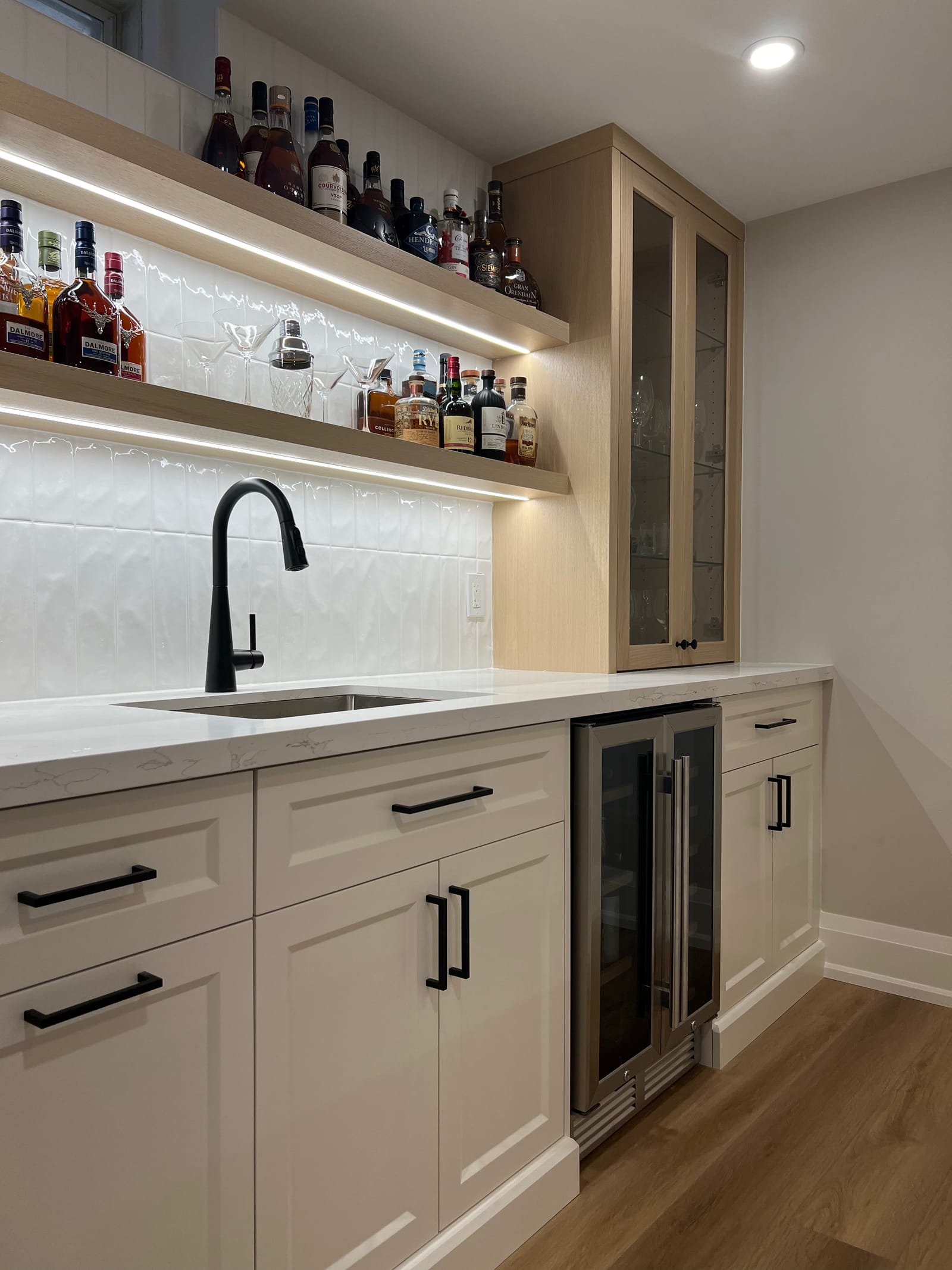 A bar run with a black faucet, a wine fridge, lit floating shelves and a glass-front oak cabinet