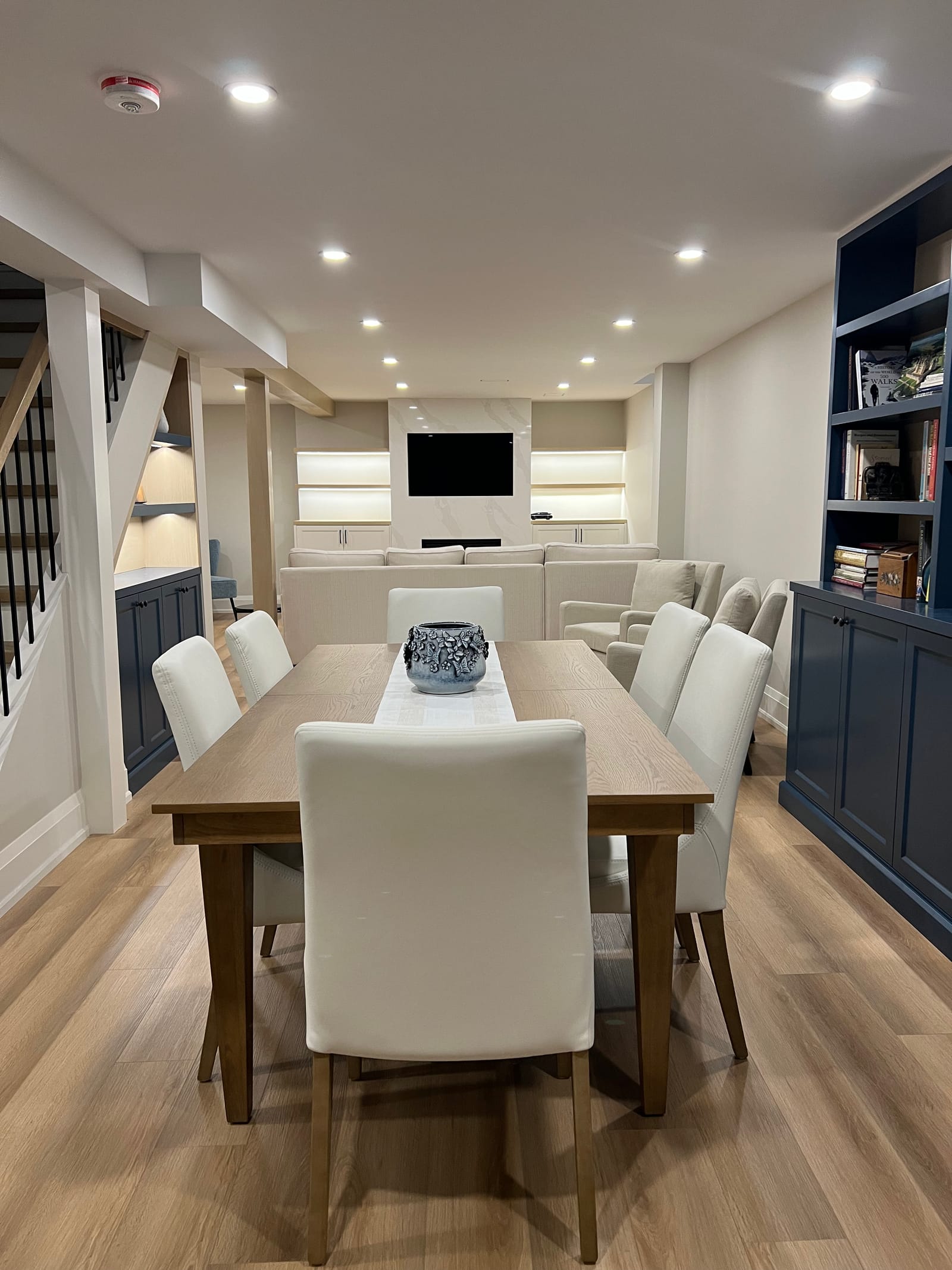A finished basement with a long dining table, a navy built-in bookcase and a media wall with a fireplace at the far end