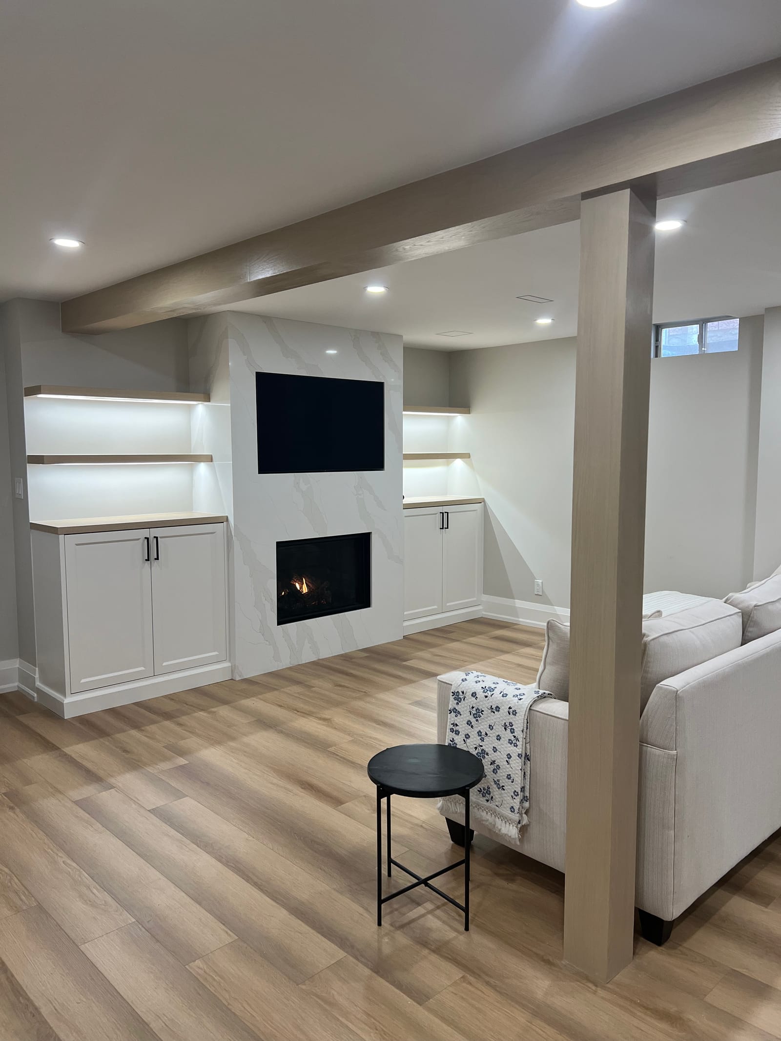 A basement media wall in white veined stone with a fireplace and TV, between cabinets and lit floating shelves