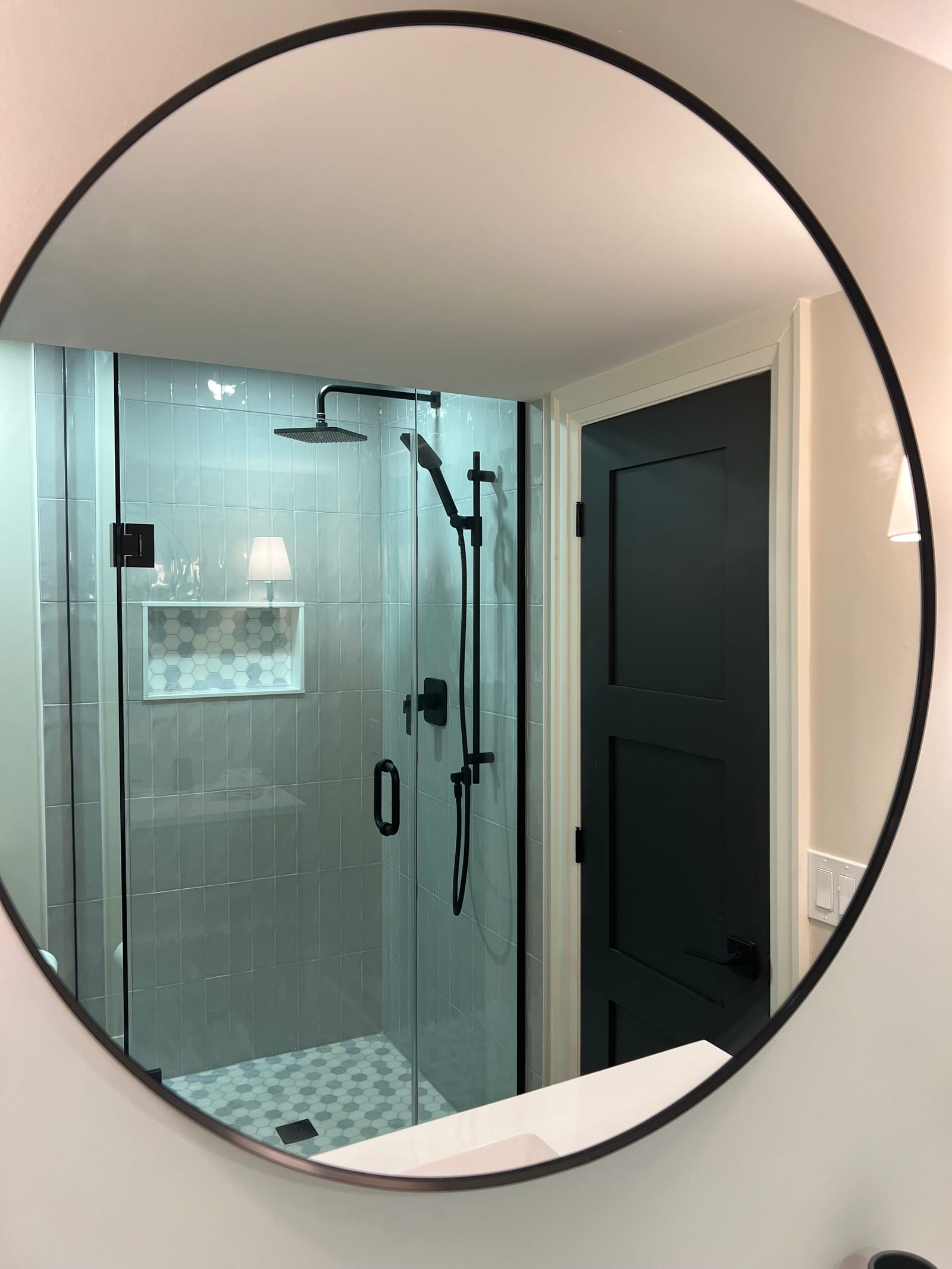 A glass shower with grey vertical tile and a hexagon niche, seen in a round mirror