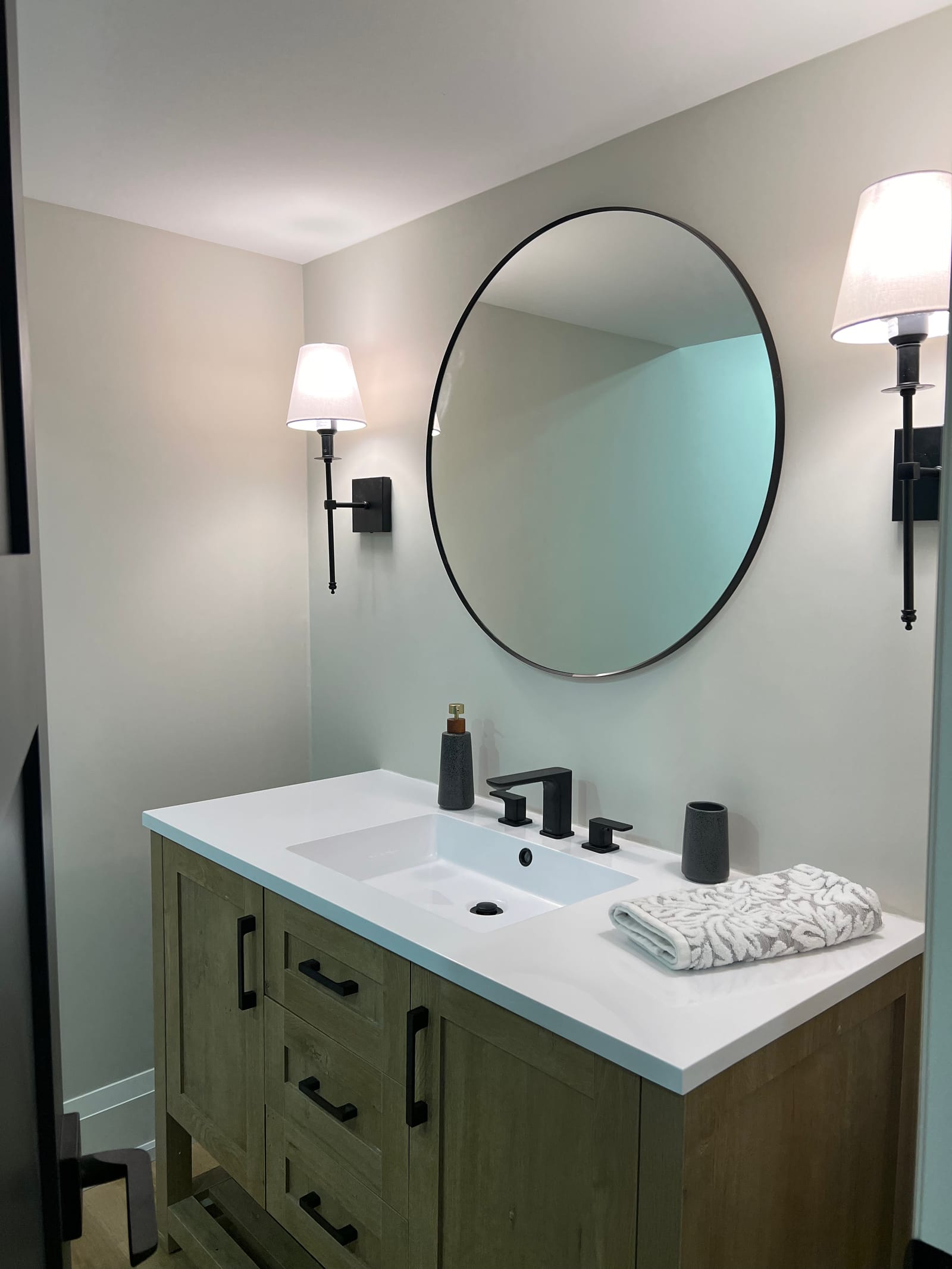 A wood vanity with a white top, black faucet, round mirror and two shaded sconces