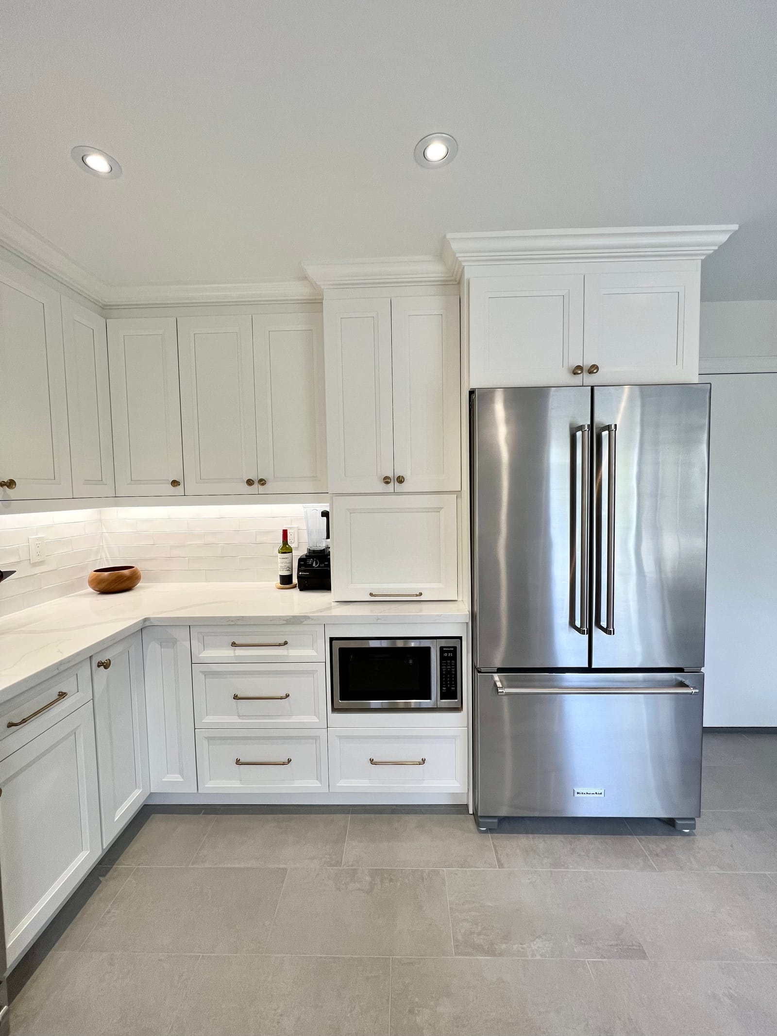 A white L-shaped kitchen corner with a built-in microwave and a stainless fridge under a tall cabinet