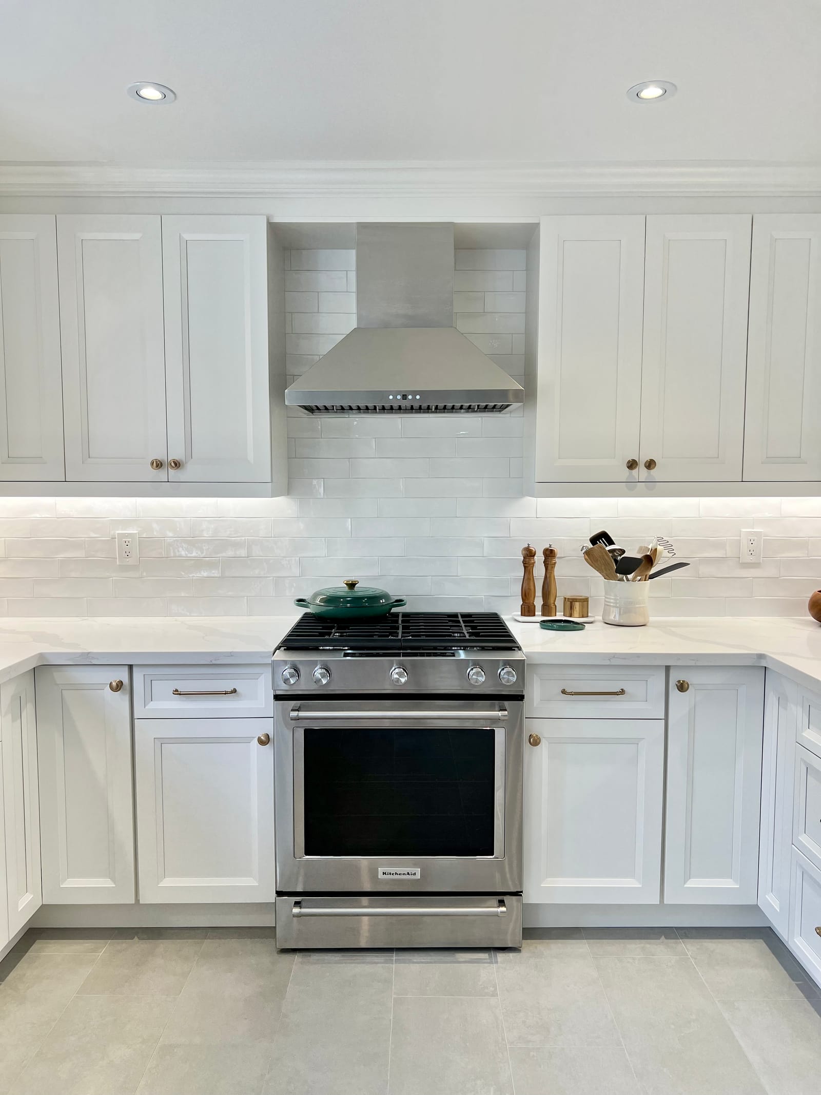 A gas range under a stainless hood between white shaker cabinets and a glossy white tile backsplash