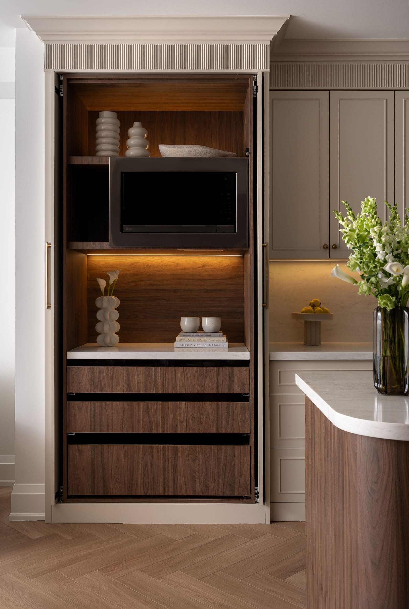 A tall cream cabinet with its doors folded back to show a walnut-lined appliance garage with a microwave, lit shelves and drawers