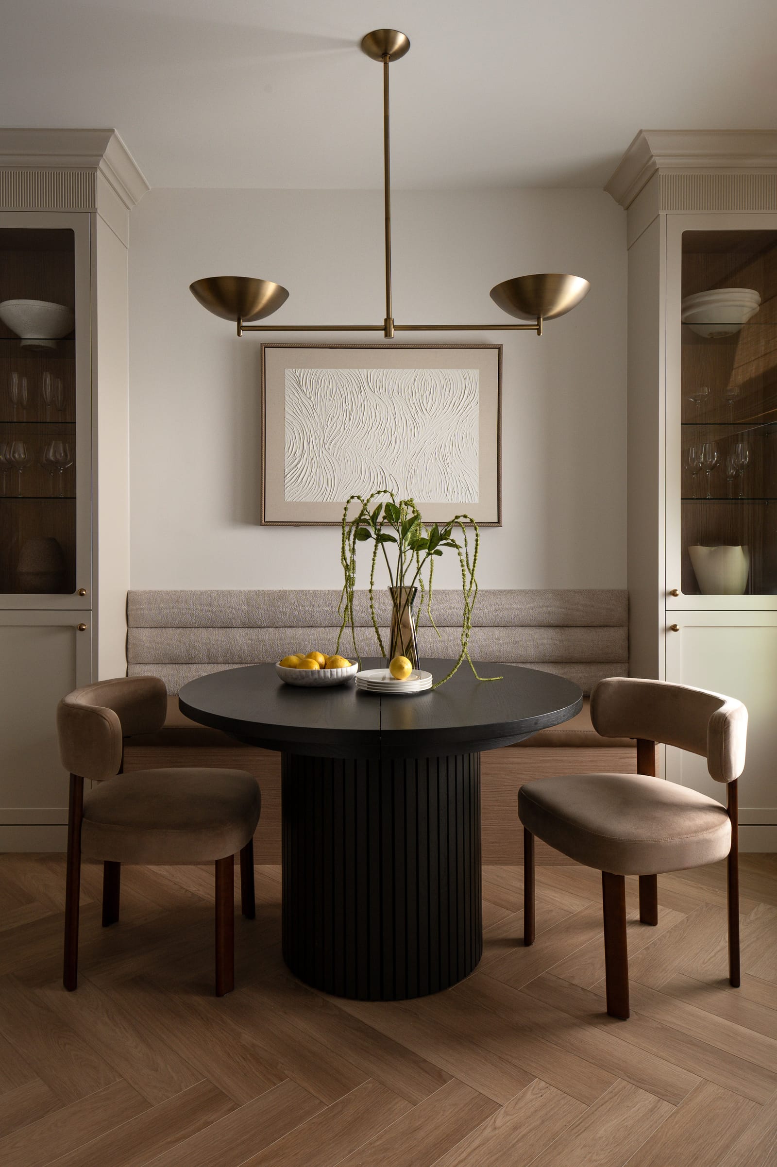 A dining nook with an upholstered banquette, a fluted black round table, two velvet chairs and glass-front cabinets on either side