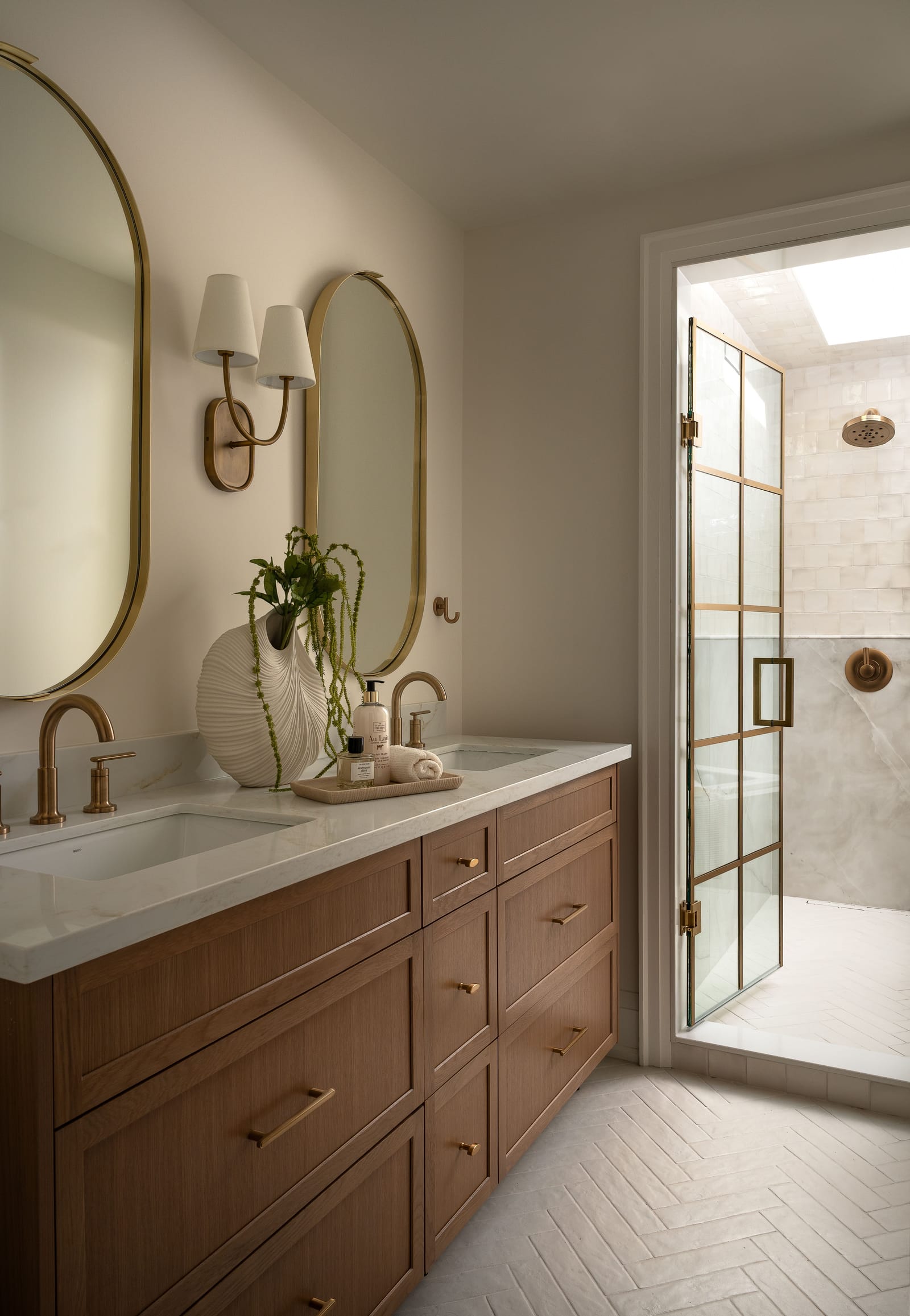 A primary ensuite with a wood double vanity, brass taps, two oval brass mirrors and a gridded glass door into a skylit wet room