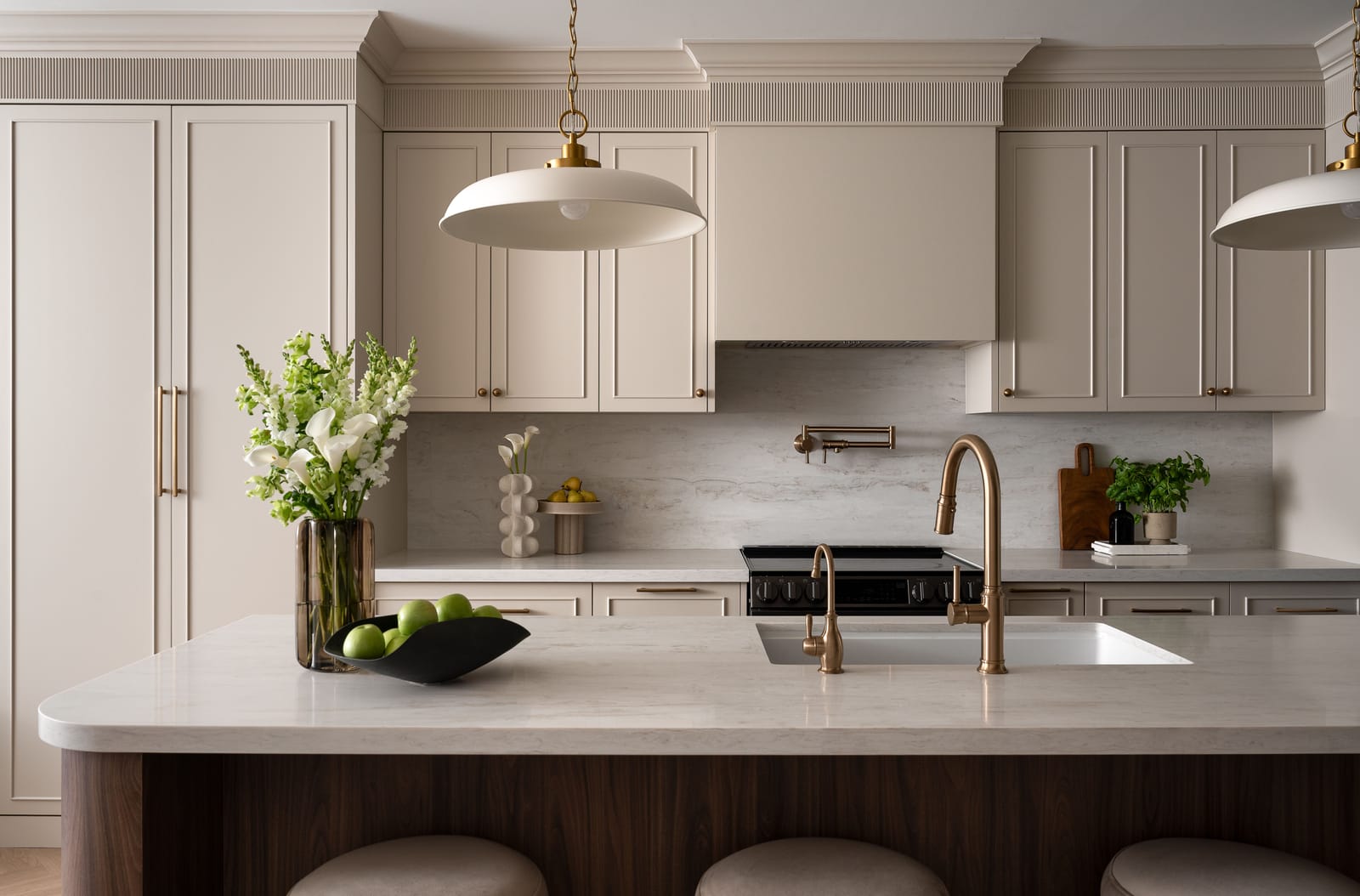 A walnut island with a rounded end and a pale stone top, in front of cream cabinets with fluted crown, a plaster hood and a brass pot filler