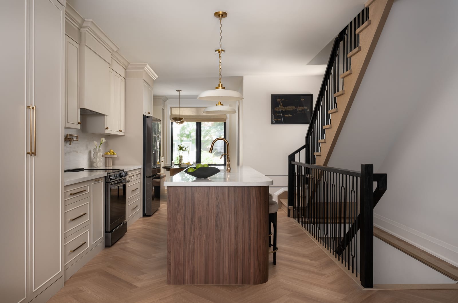 A long kitchen seen from the front of the house: cream cabinets on the left, a rounded walnut island in the middle and an open oak stair with black railings on the right
