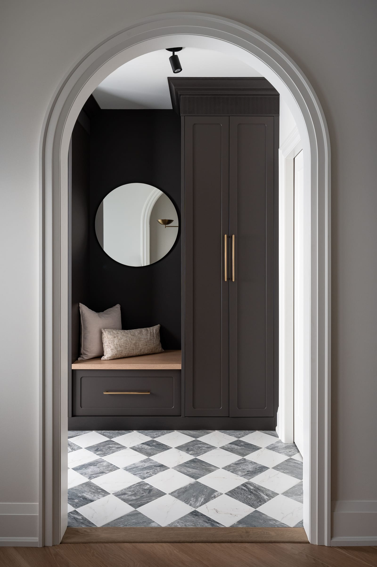 An arched doorway framing a charcoal mudroom with a bench, a tall cabinet with brass pulls, a round mirror and a grey and white checkerboard marble floor