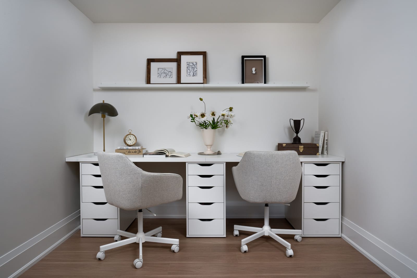 A home office with a long white desk on drawer units, two grey swivel chairs and a single floating shelf of framed prints