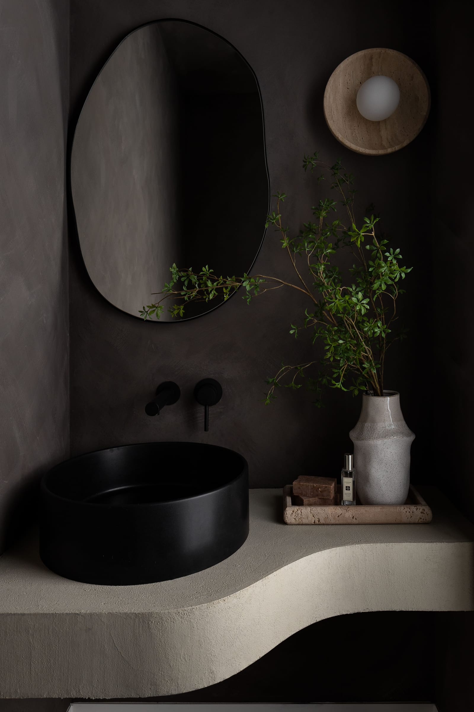 A dark micro-cement powder room with a curved floating vanity, a black vessel sink, wall-mounted black taps and an organic-shaped mirror