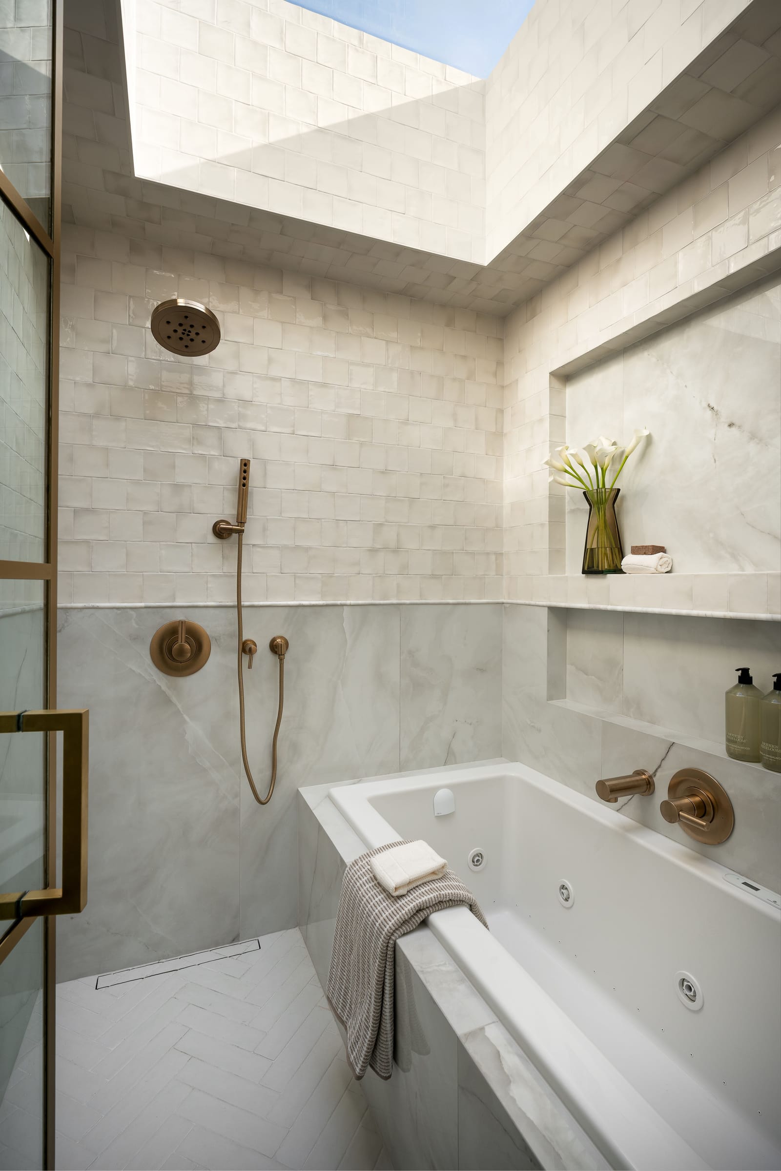A wet room lit by a skylight, with zellige-style wall tile, marble-look slabs, a jetted tub, a brass rain shower and a linear drain