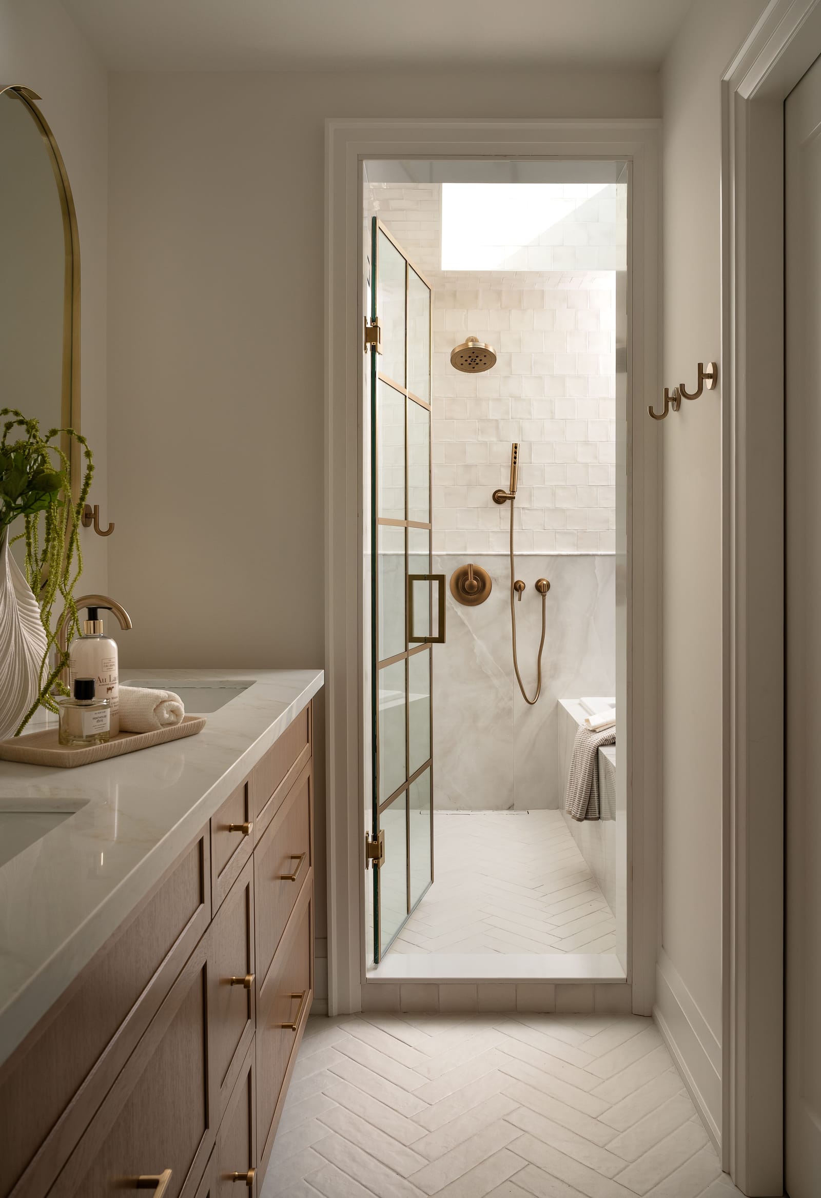 Looking from the ensuite through a gridded brass glass door into the wet room, with a brass rain shower under the skylight and a herringbone tile floor