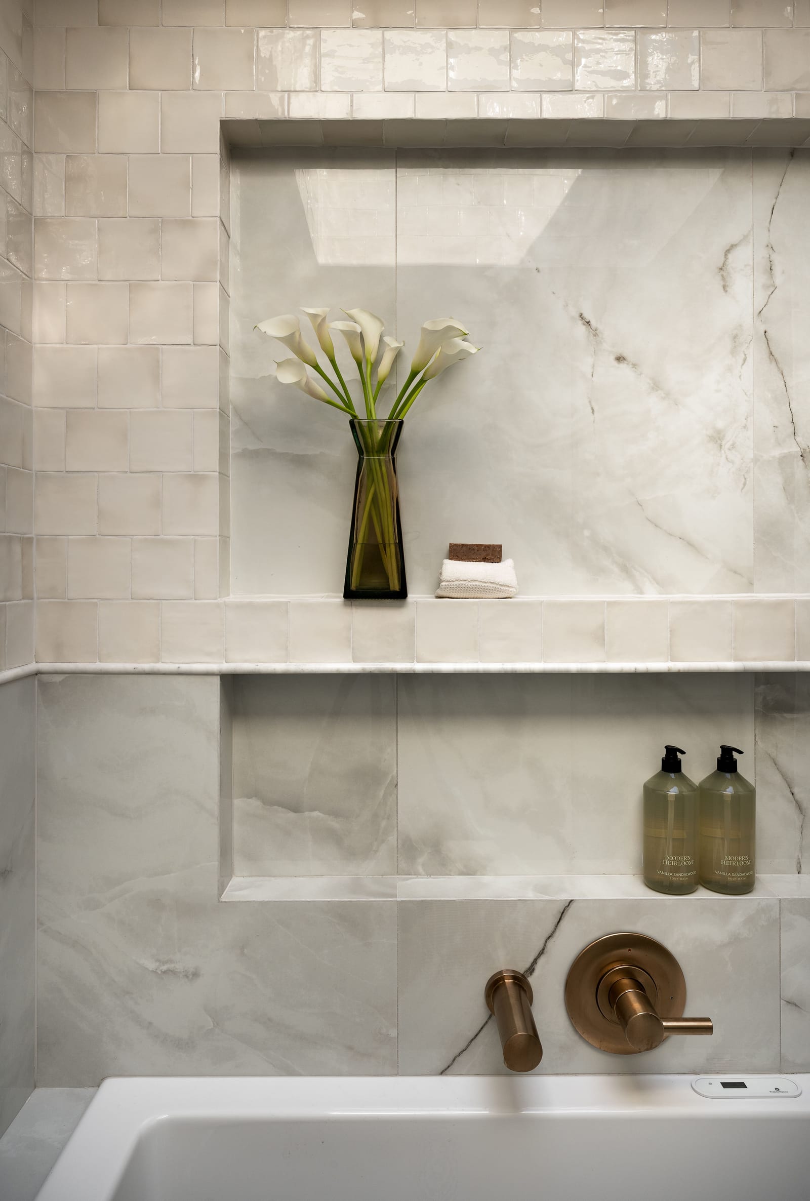 Glossy zellige-style tile over marble-look slabs in the wet room, with two long niches holding calla lilies and body wash above a brass tub filler