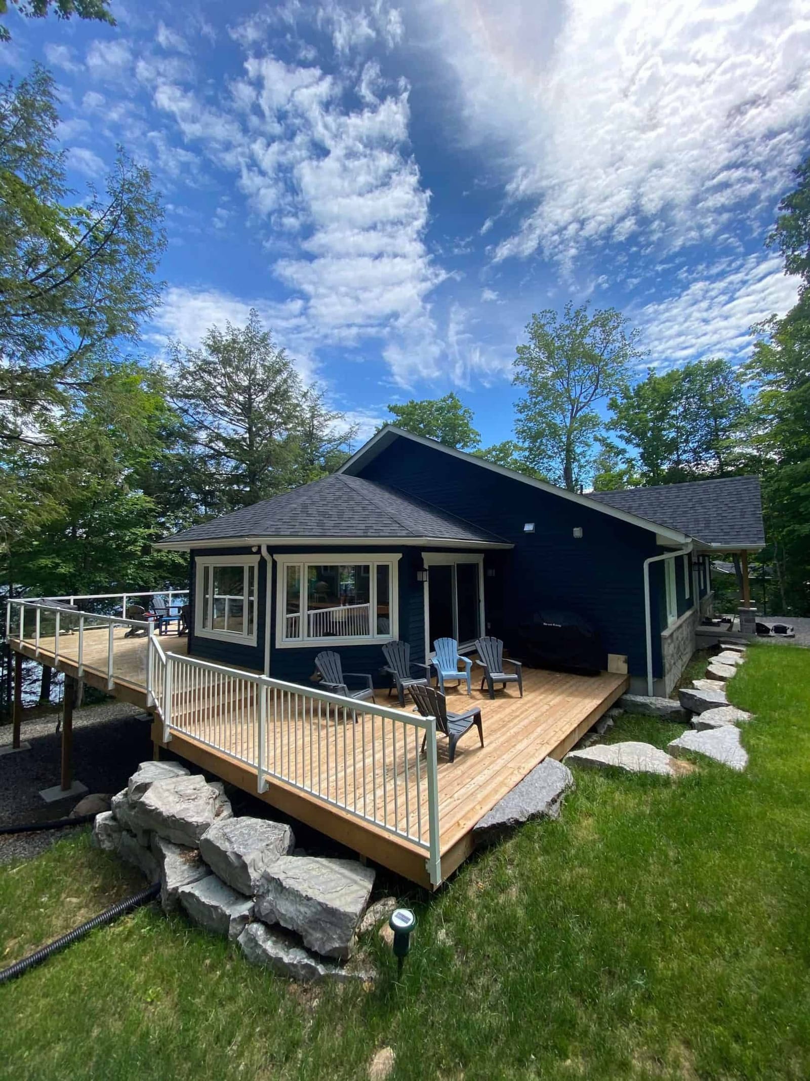 A wide cedar deck with white railings wrapping a navy house on a rock ledge, among tall trees