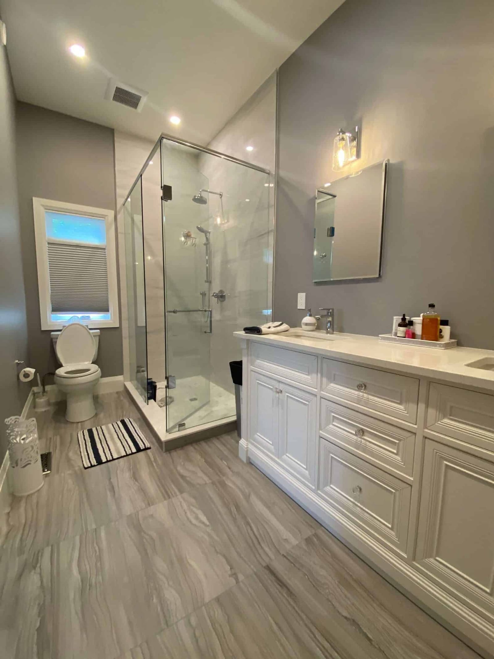 A bathroom with a long white vanity, a glass corner shower and a large-format grey tile floor