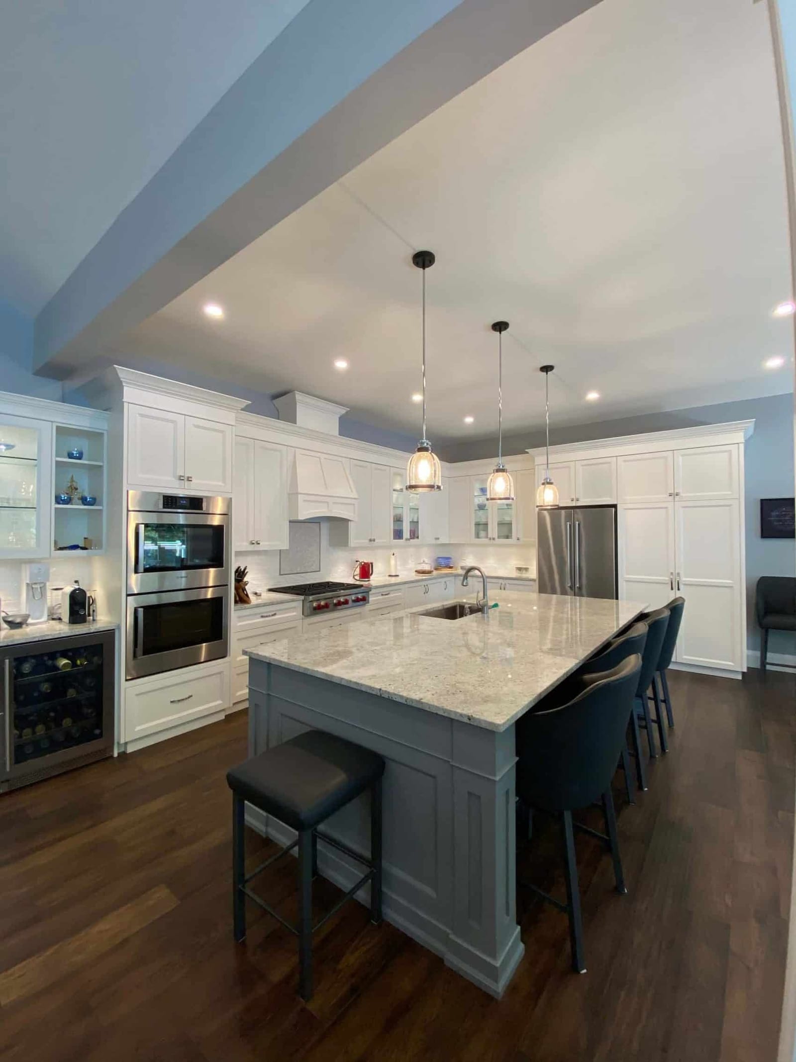 A white kitchen with a grey island under three glass pendants, double wall ovens and a wine fridge