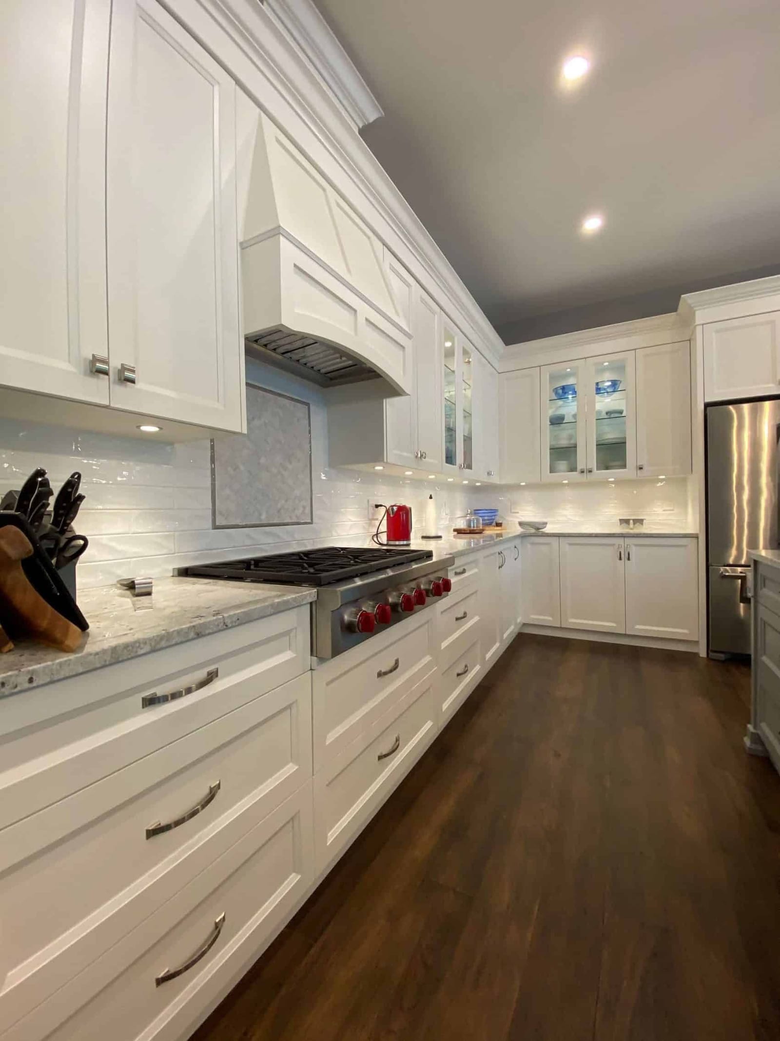 A white kitchen run with a gas range, a white hood and glass-front upper cabinets