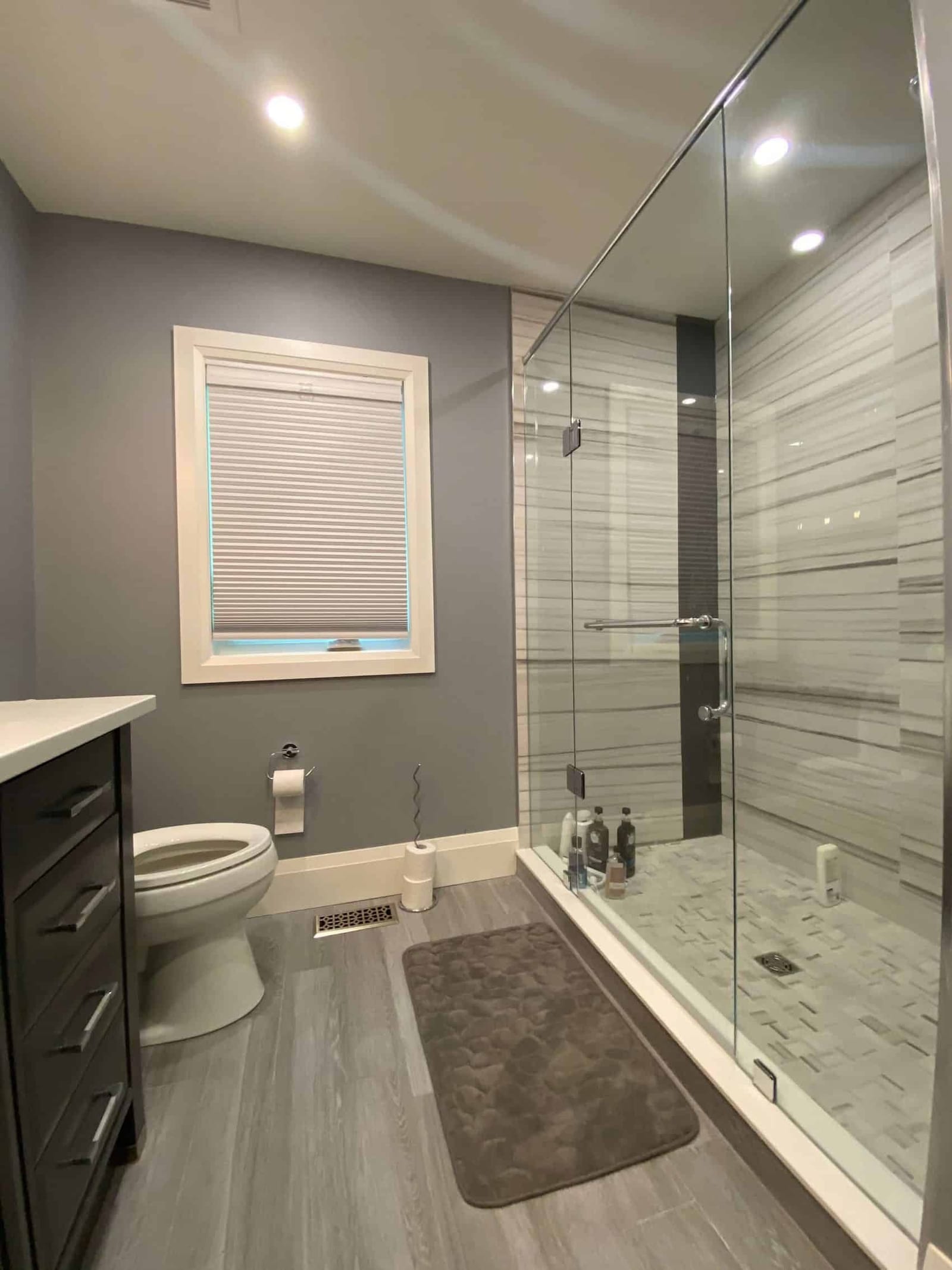 A walk-in glass shower in striped grey stone tile beside a dark vanity