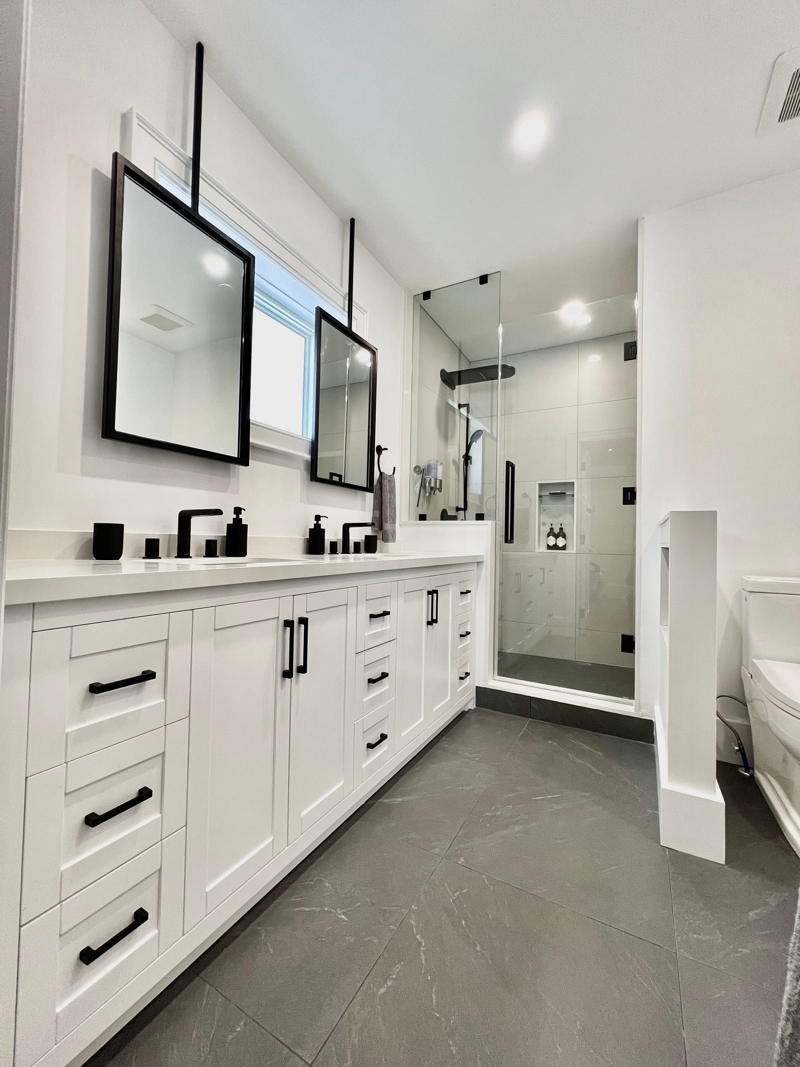 A white double vanity with black hardware and ceiling-hung black mirrors, a glass shower and a charcoal floor
