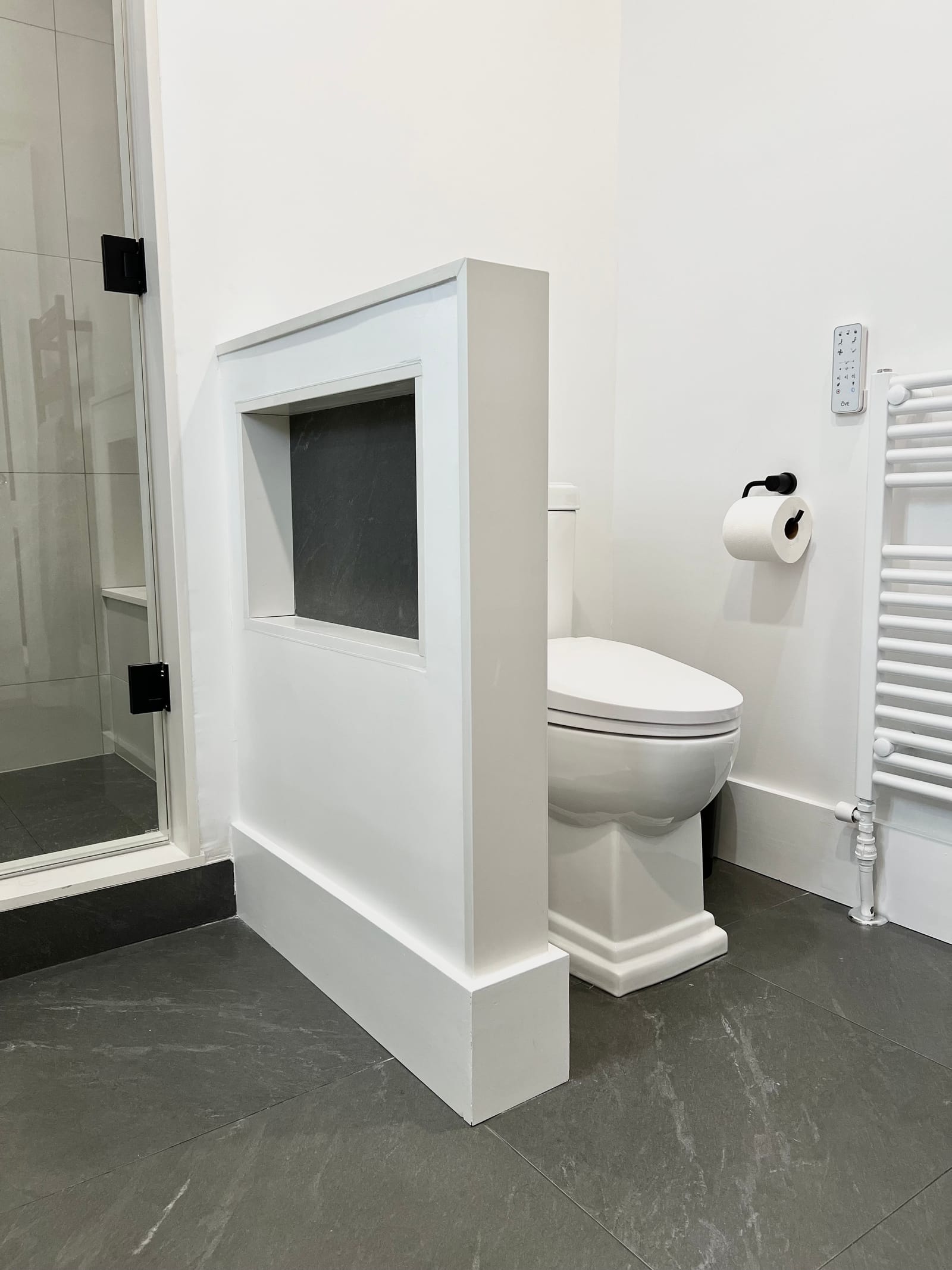 A half wall with a dark niche screening the toilet, beside the glass shower