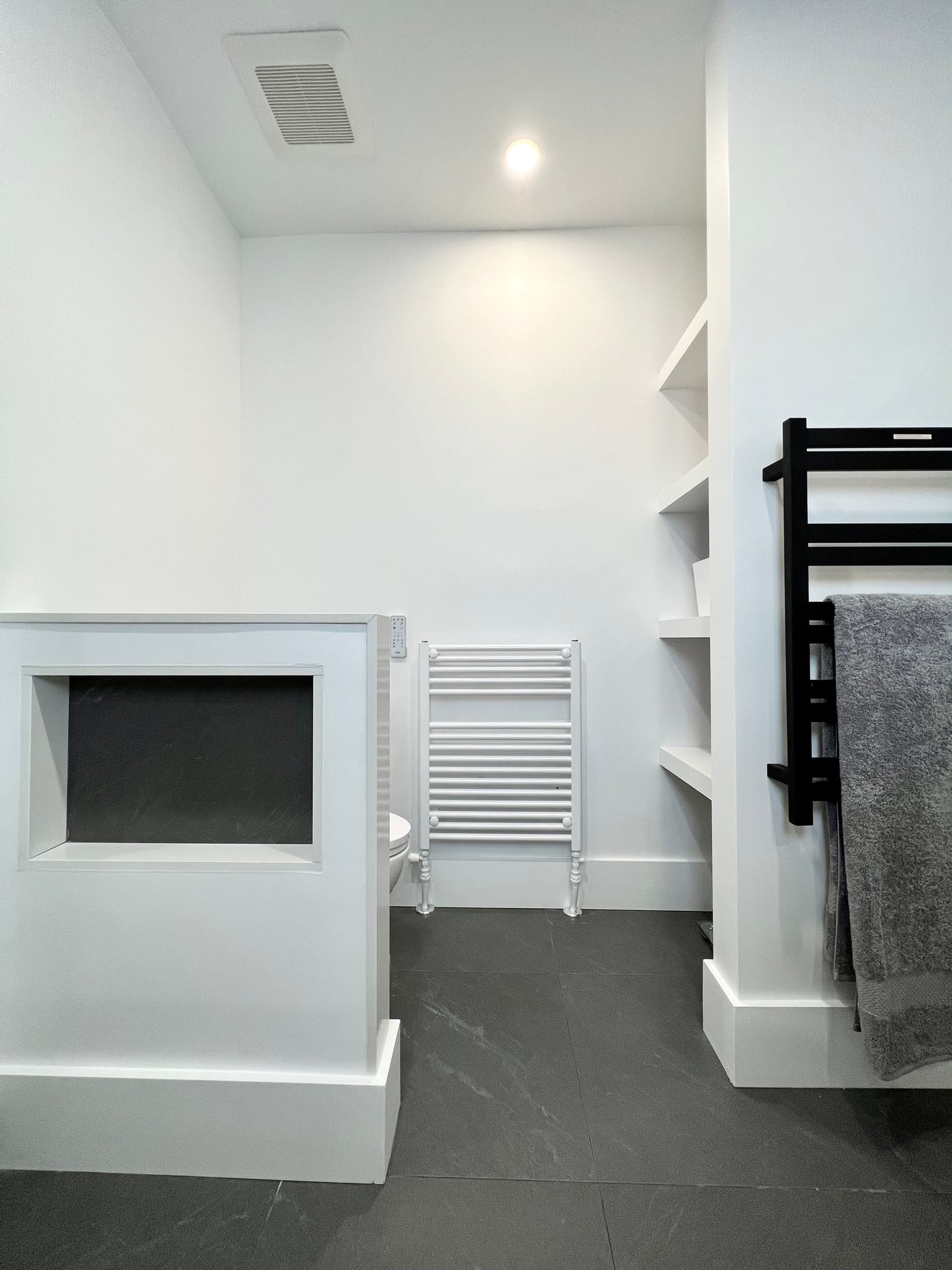 A half wall with a niche, black and white towel warmers and open shelves on a charcoal floor